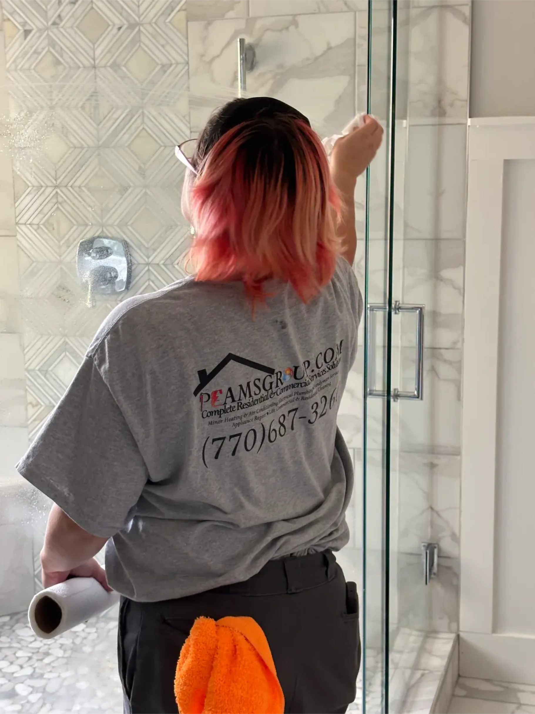 Person painting a bathroom wall, wearing a gray shirt and black shorts, with pink hair.