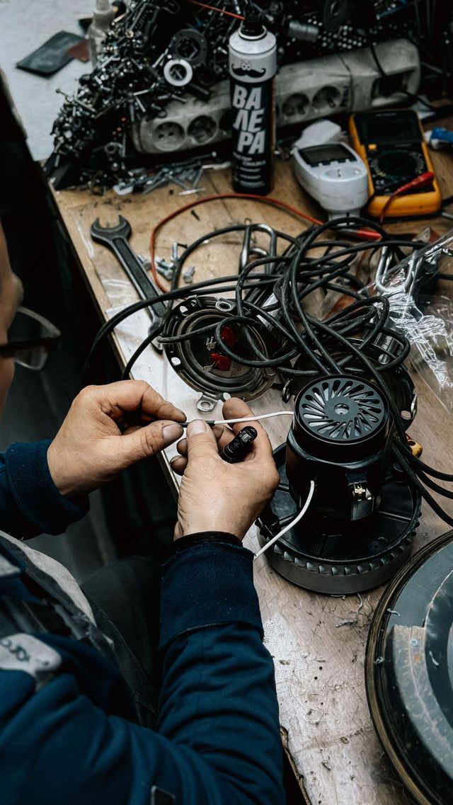 Person assembling tangled cables and electronics on a cluttered workbench with tools and parts