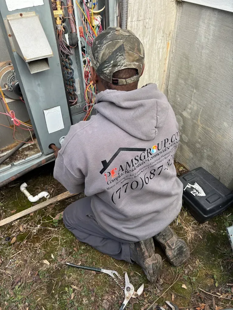 Person kneeling beside electrical equipment outdoors, wearing a gray hoodie and camo cap, working on wiring.