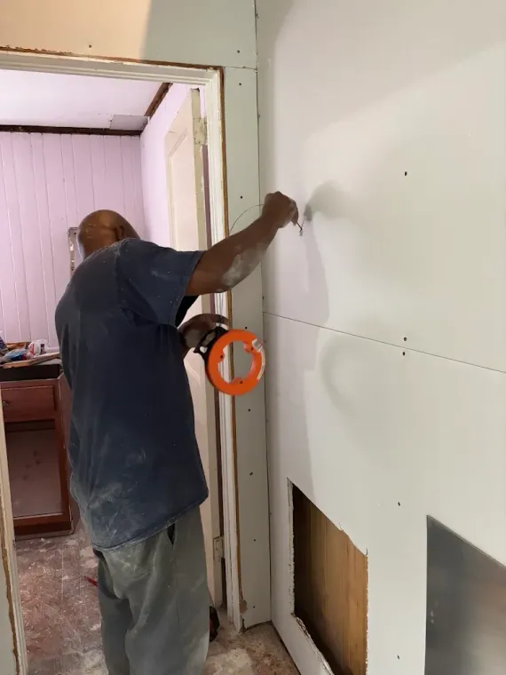 Worker installing a white wall panel beside a doorway, holding a tool in an unfinished room.
