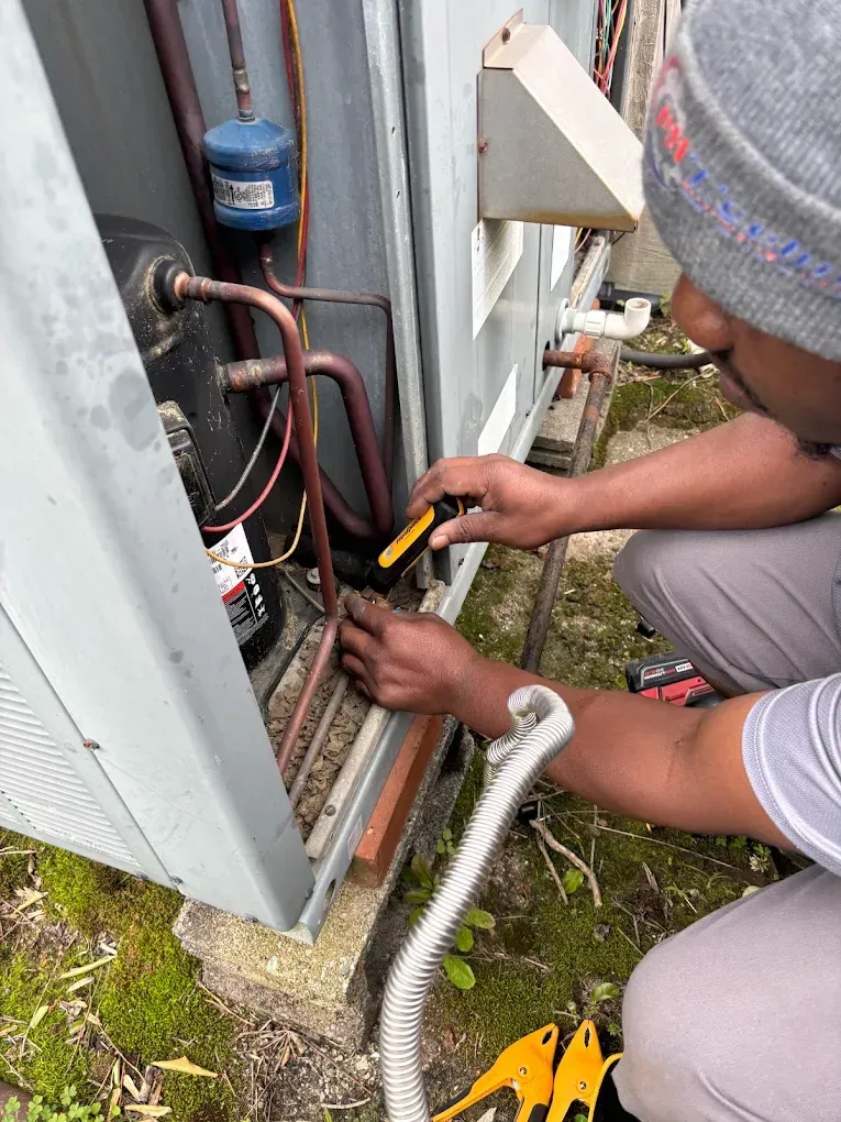 Technician repairing wiring on an outdoor AC unit, using tools beside exposed pipes and panels.