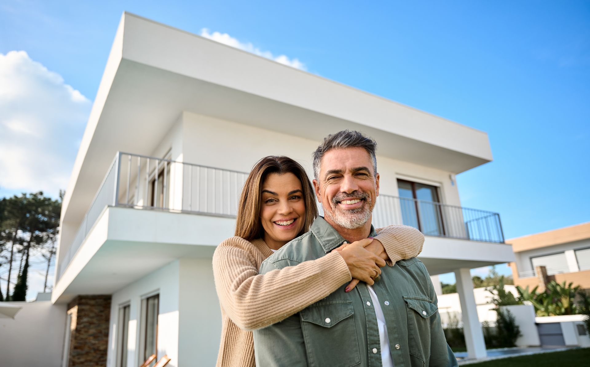 Smiling couple embracing in front of a modern white house with balconies under a bright sky