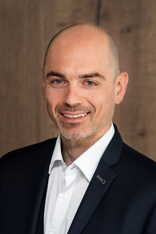 Mitarbeiter der cts in einem dunklen Anzug, lächelt vor einem hölzernen Hintergrund.