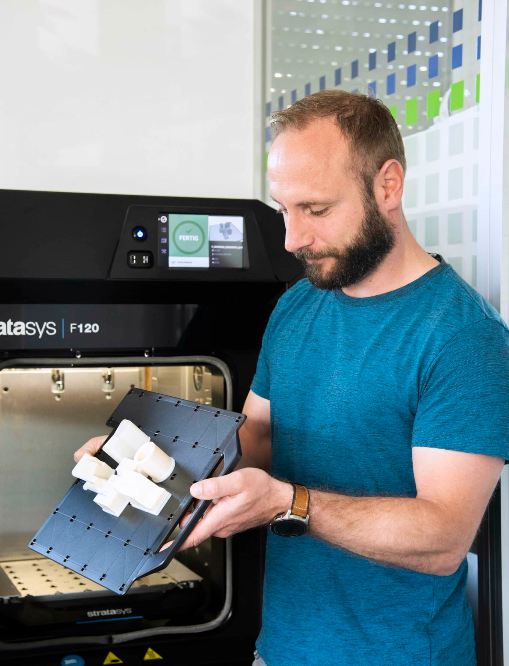 Ein Mann in einem blauen Hemd hält ein Stück Plastik vor einen 3D-Drucker.