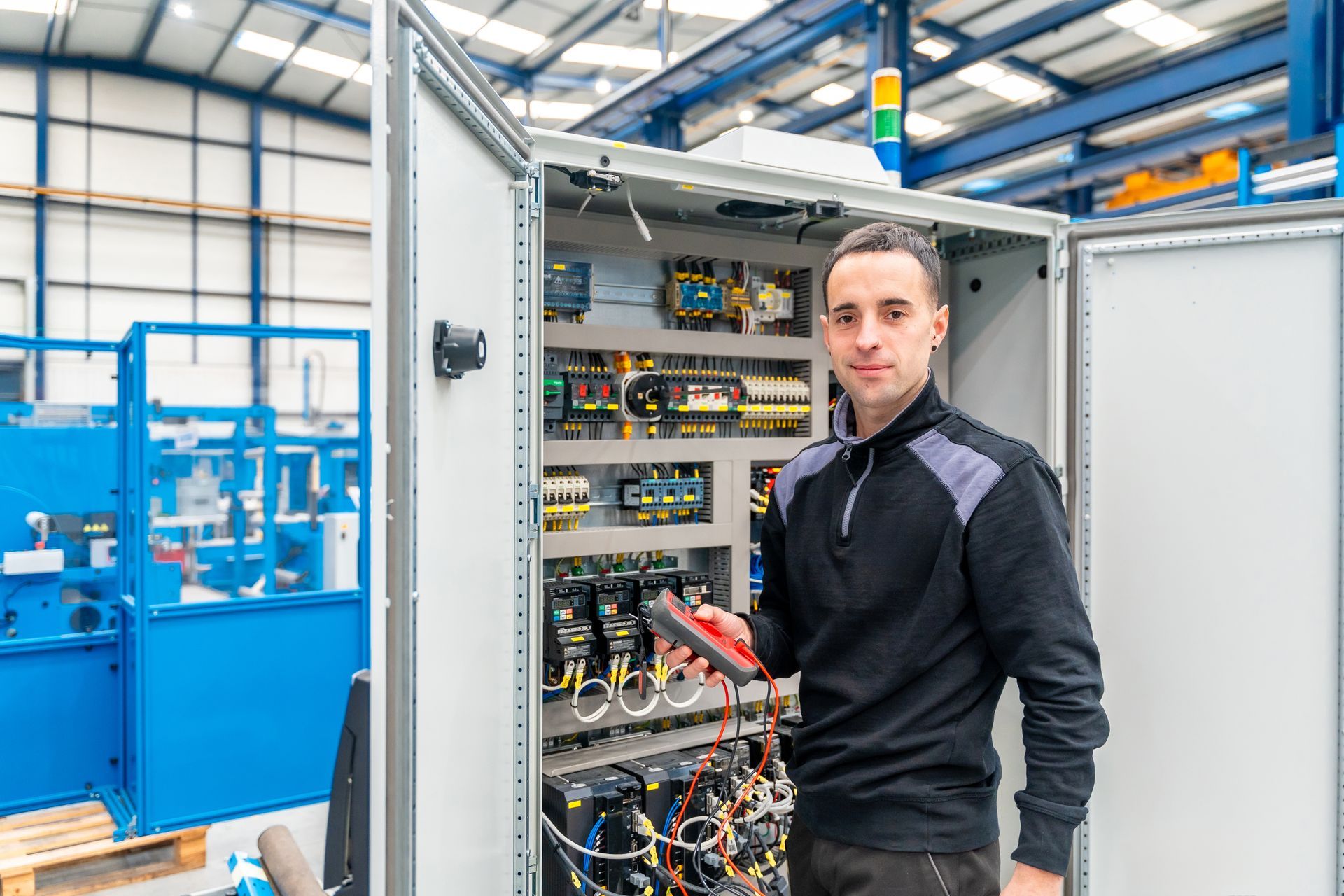 Man in black shirt holding a meter, working on electrical panel in a factory.