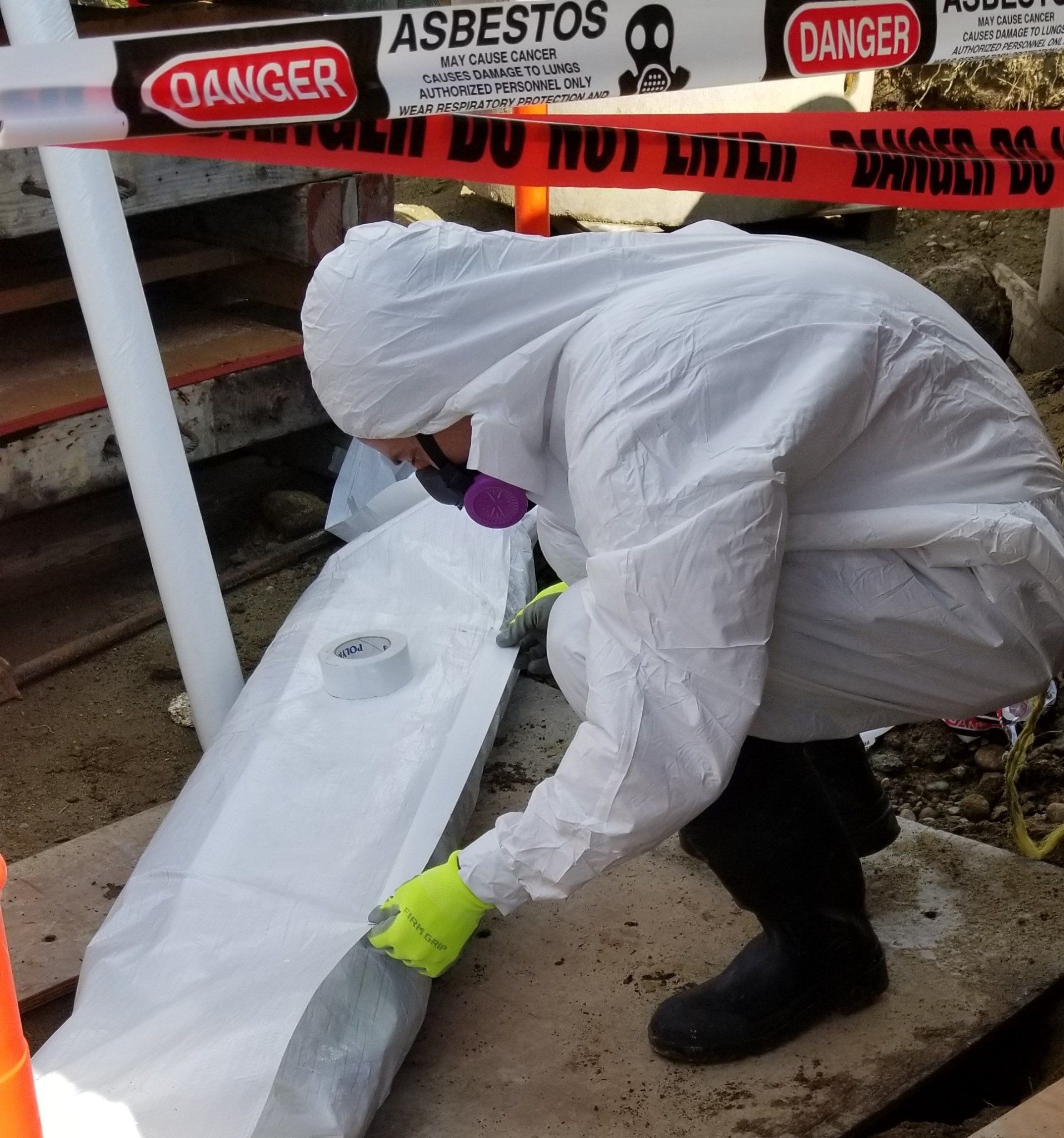Worker in white protective suit handling a long wrapped object near asbestos warning tape.