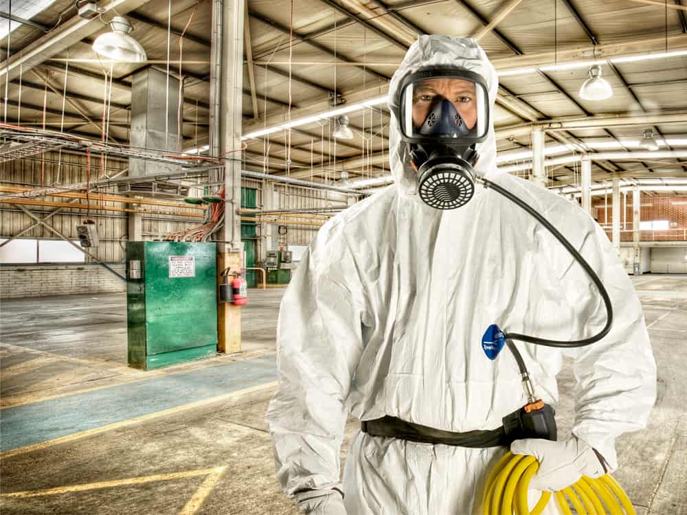 Person in hazmat suit and respirator standing in an industrial warehouse.