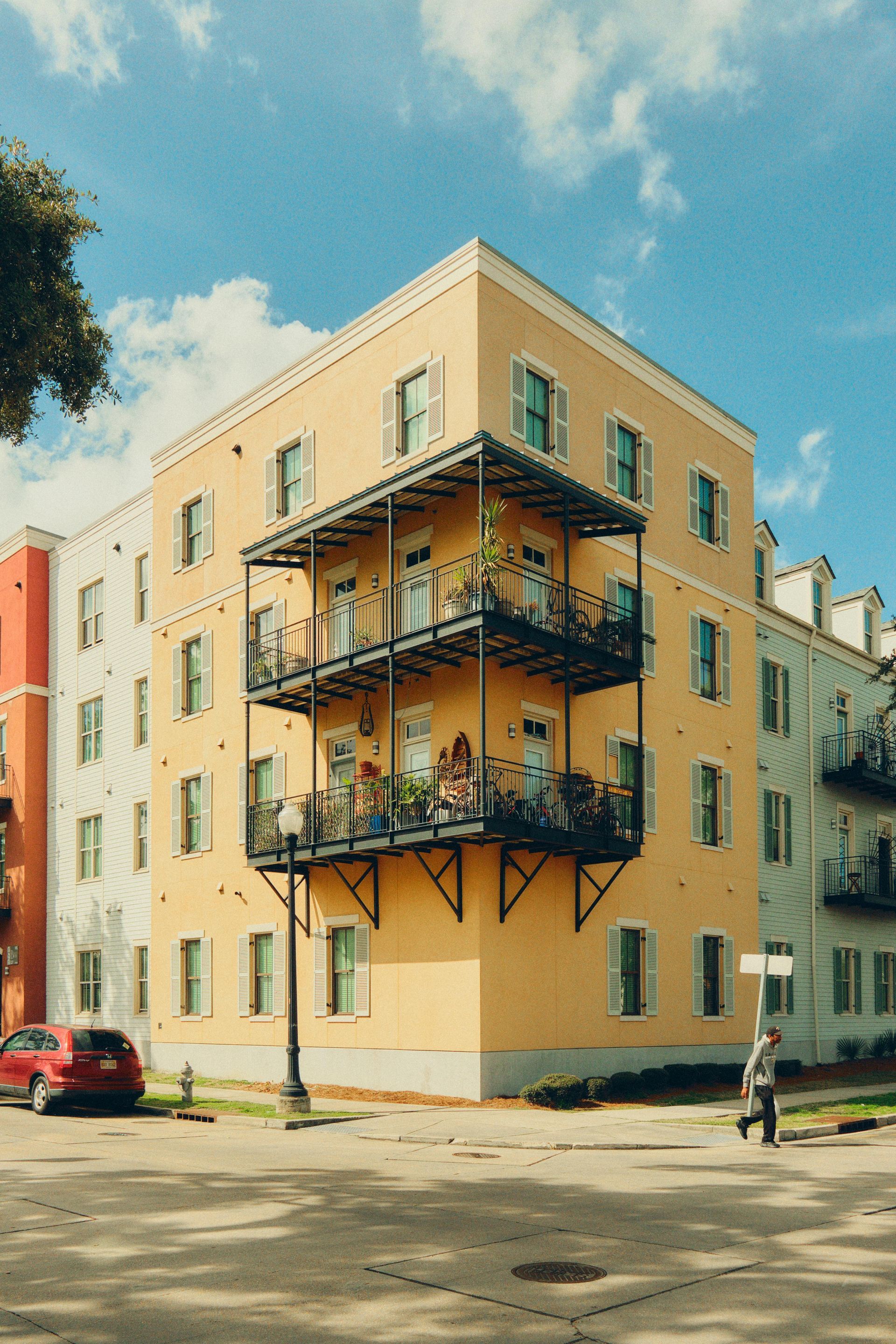 Yellow apartment building with black balconies, car, and pedestrian on street.
