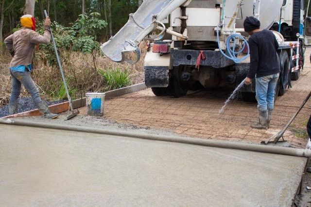 Workers pouring concrete from truck onto roadbed. One sprays water, another levels with a broom.