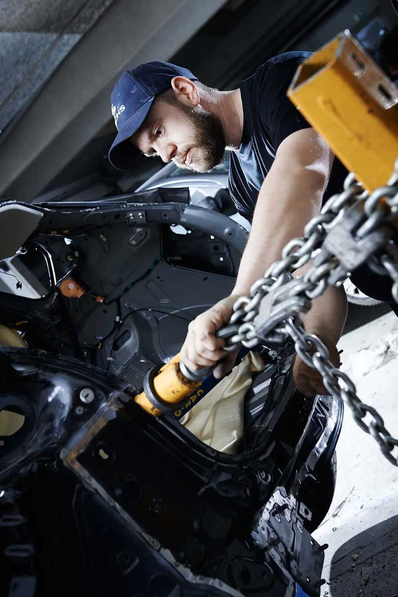 En person med en blå kasket bruger en kæde og et værktøj til at trække i metalrammen på et køretøj under en bilreparation.