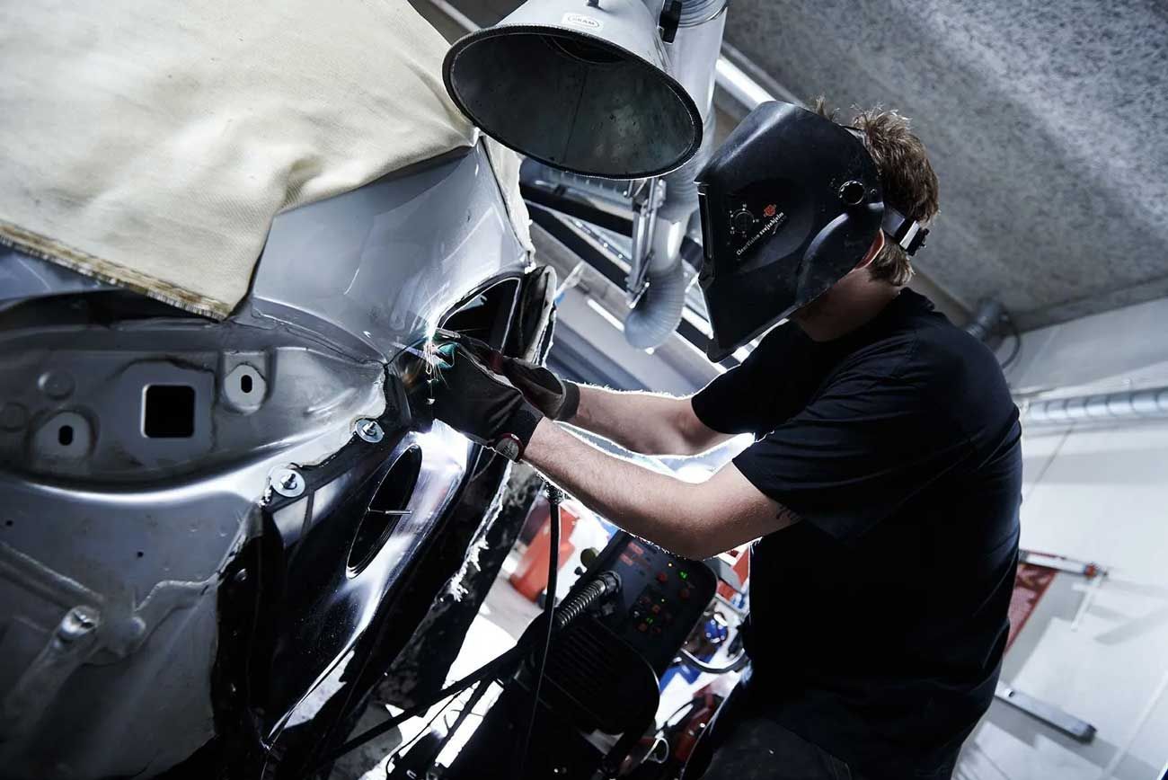 En person iført svejsehjelm og handsker bruger et værktøj på en bils metalramme i et autoværksted.