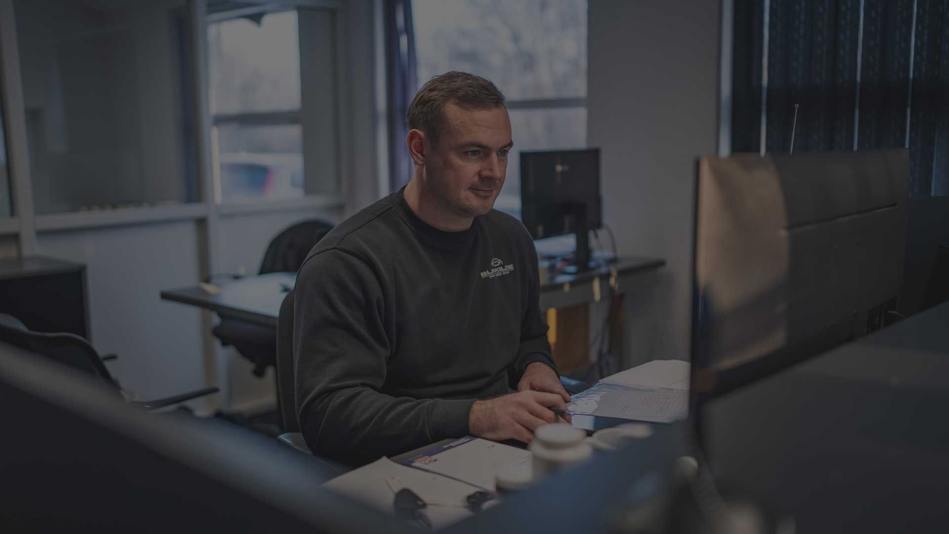 En person sidder ved et skrivebord på et kontor, arbejder på en computer og gennemgår papirer.