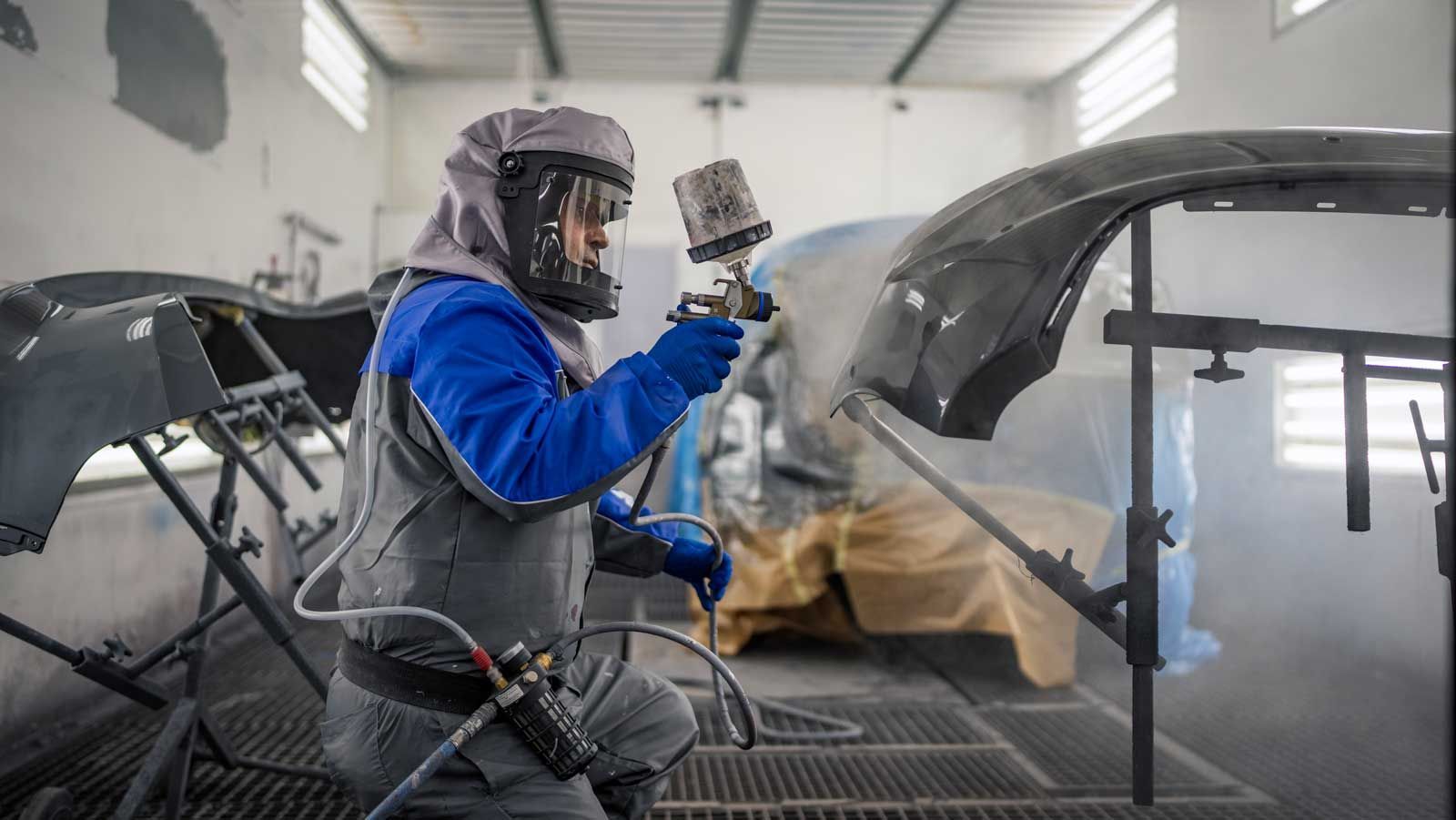 En arbejder i beskyttelsesudstyr og åndedrætsværn sprøjter maling på en bilkofanger inde i en værkstedsboks.