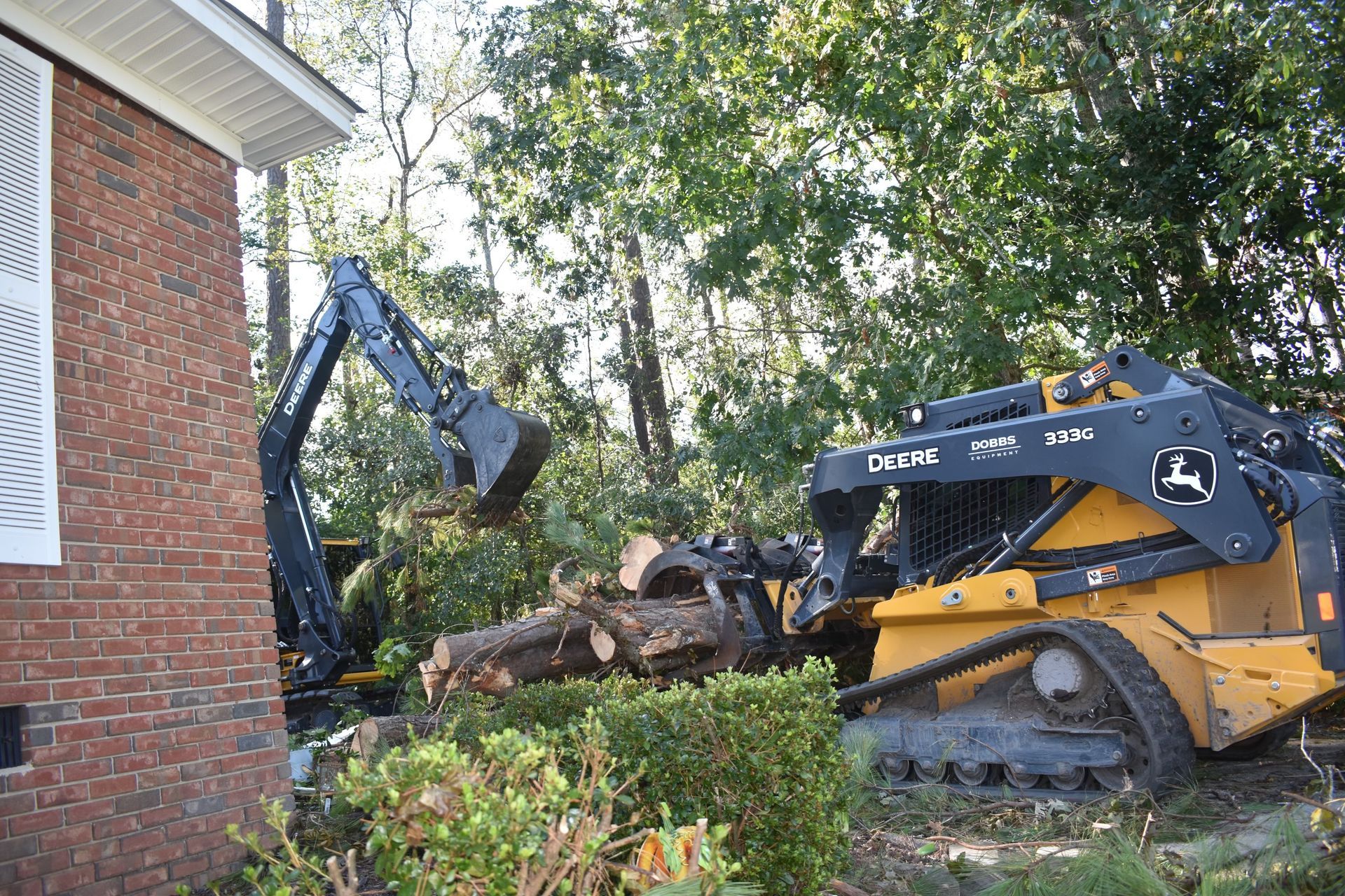 A yellow John Deere tracked skid steer with a grapple attachment moves logs near a brick building and green trees.