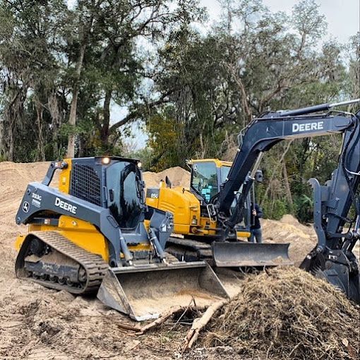 A yellow and black John Deere skid steer loader and a track-mounted excavator sit on a dirt lot near trees.