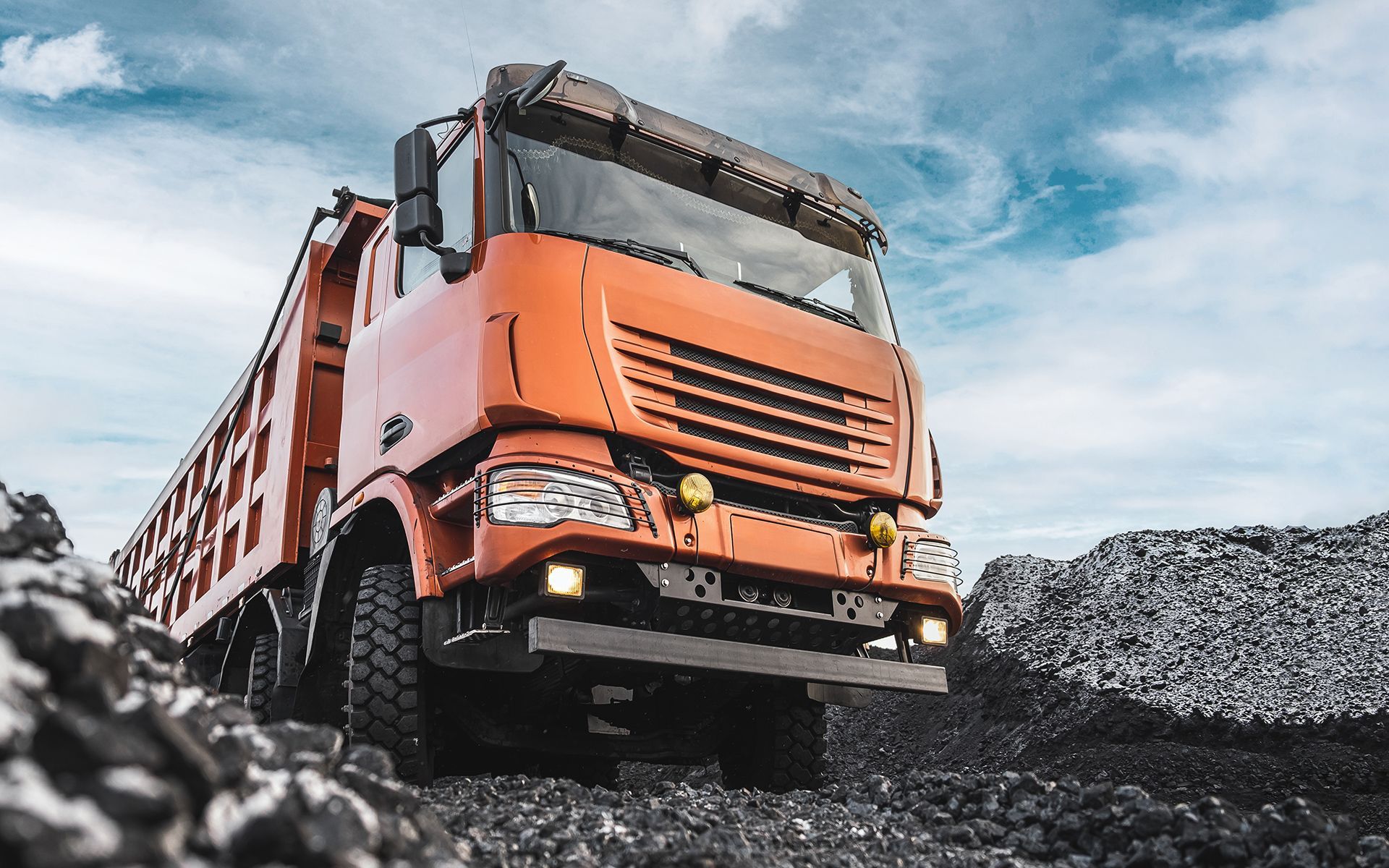 Orange dump truck driving on rocks against a cloudy sky.