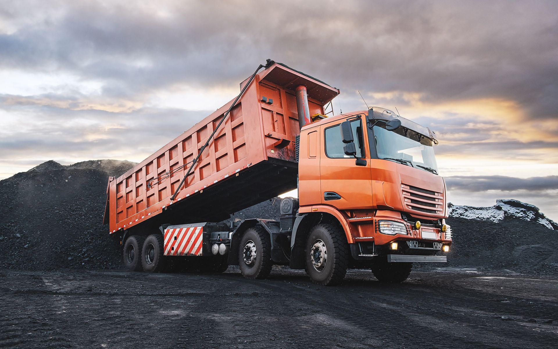 Orange dump truck unloading material onto a dark pile under a cloudy sky.