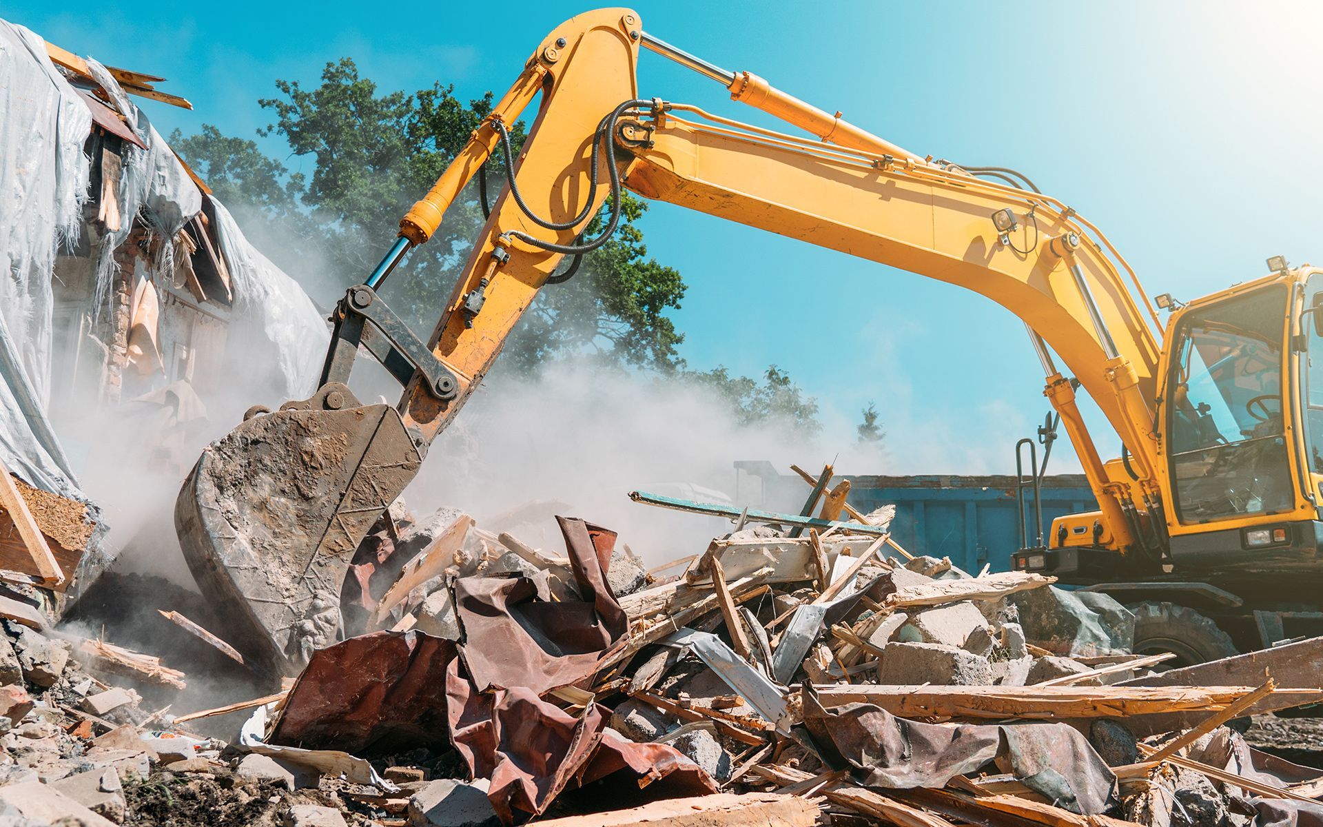 Yellow excavator demolishing a building, kicking up dust under a blue sky.