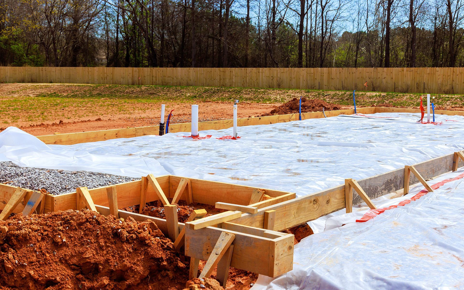 Construction site with wooden forms for a foundation, white plastic, and pipes on dirt.