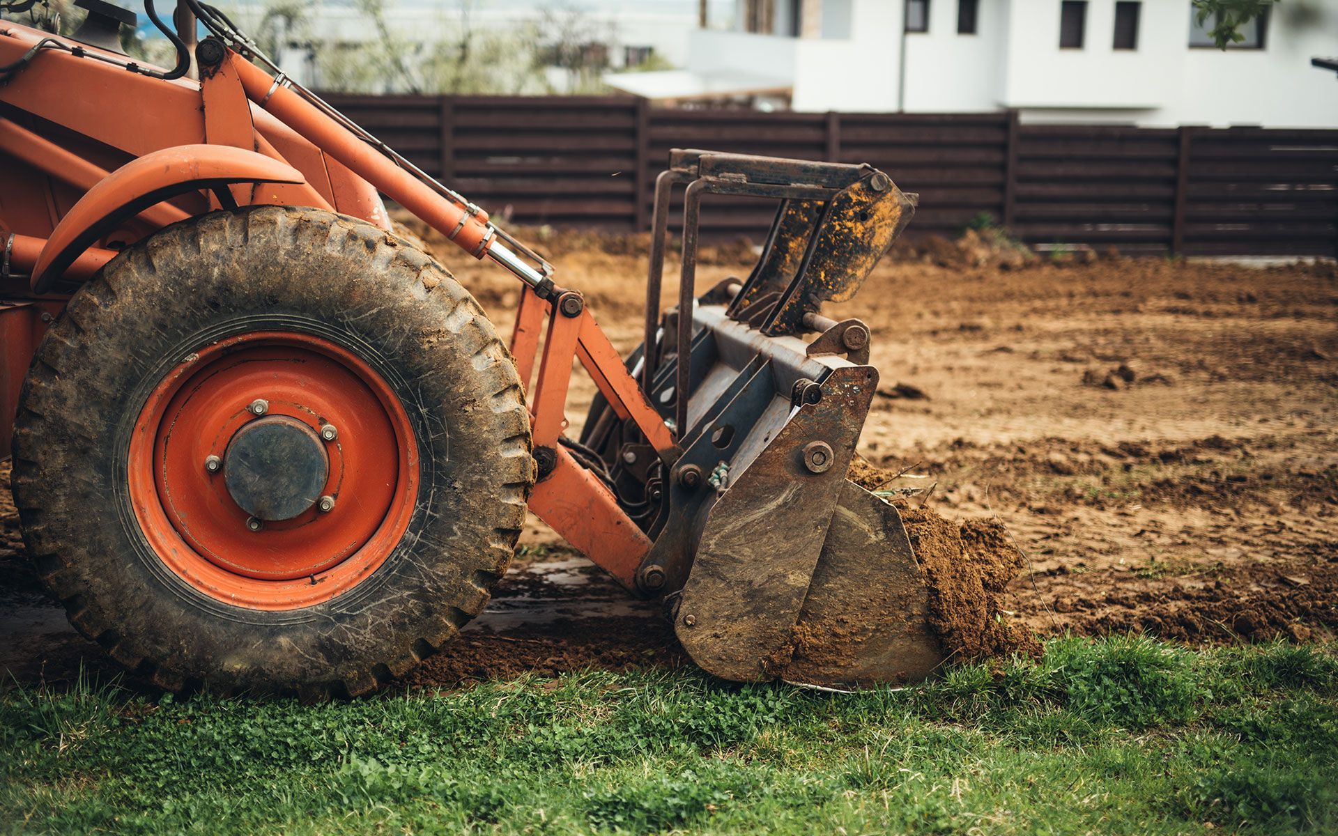 Orange tractor with bucket moving dirt on a brown field, near green grass and a wooden fence.