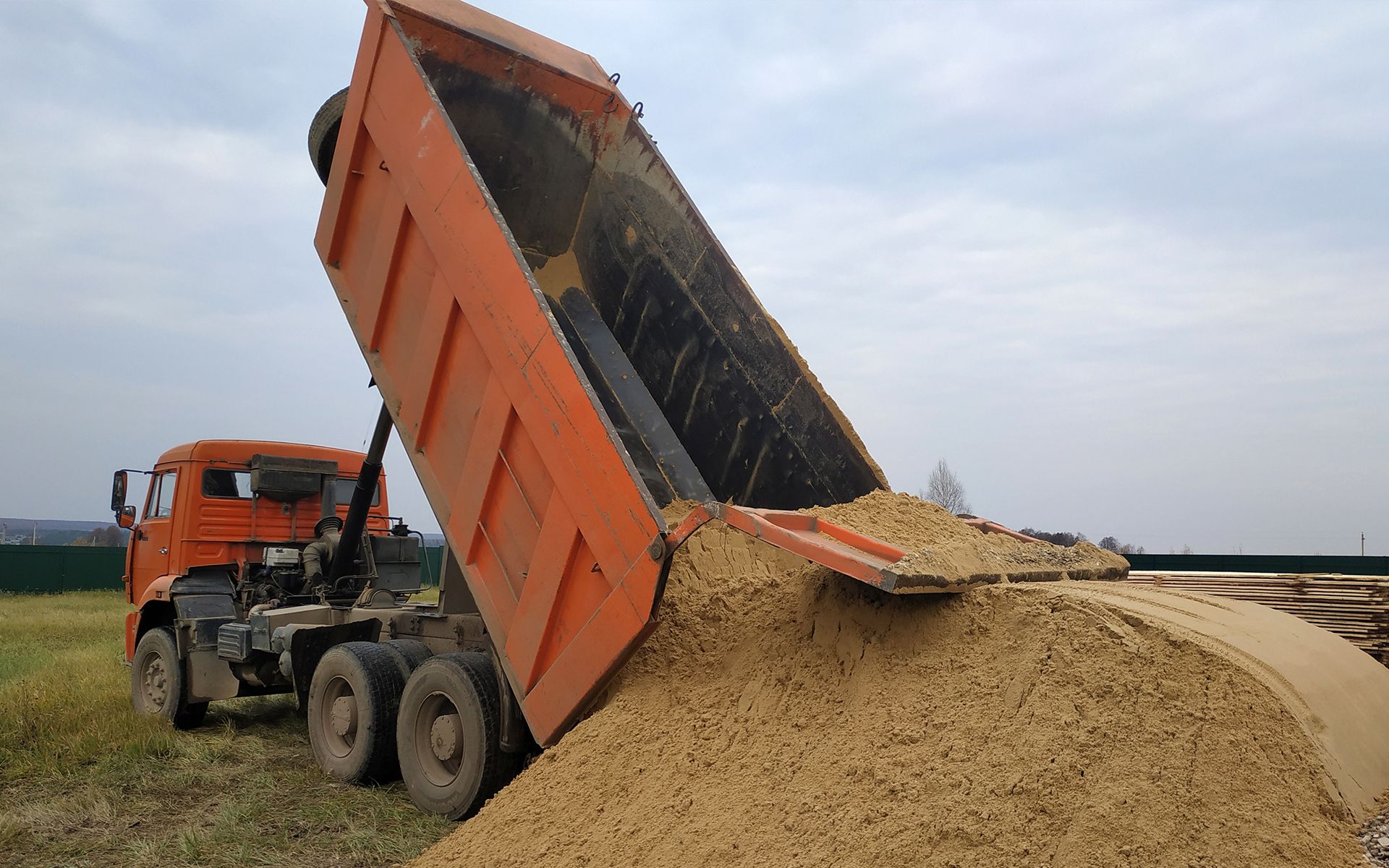Orange dump truck dumping load of brown material on a grassy field.