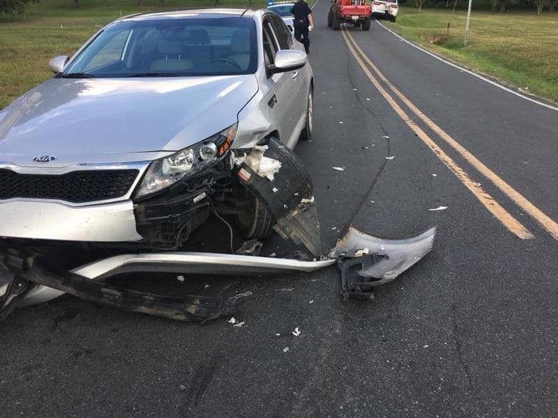 Silver Kia sedan with significant front-end damage on a road; police vehicles in the background.