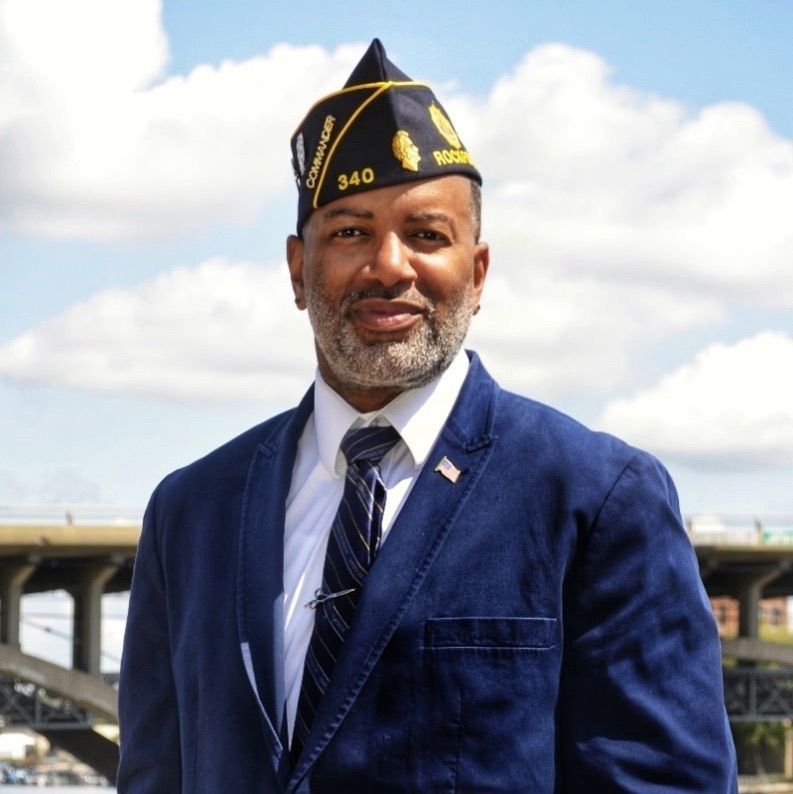 Man in blue suit and veteran's cap, smiling, outdoors against a cloudy sky and bridge.