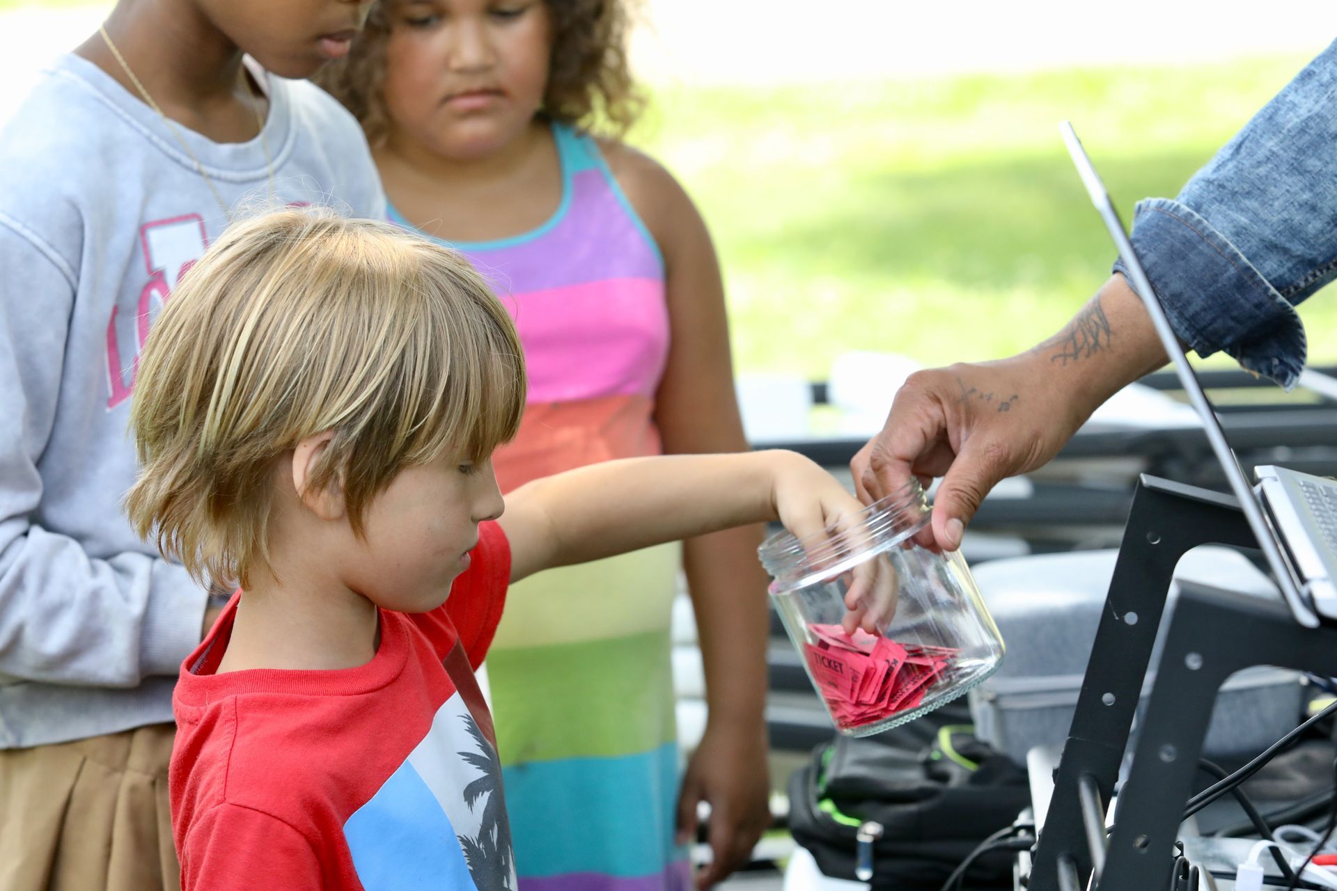 Boy in red shirt reaching into a jar held by an adult; two children watch nearby. Outdoors, sunny.
