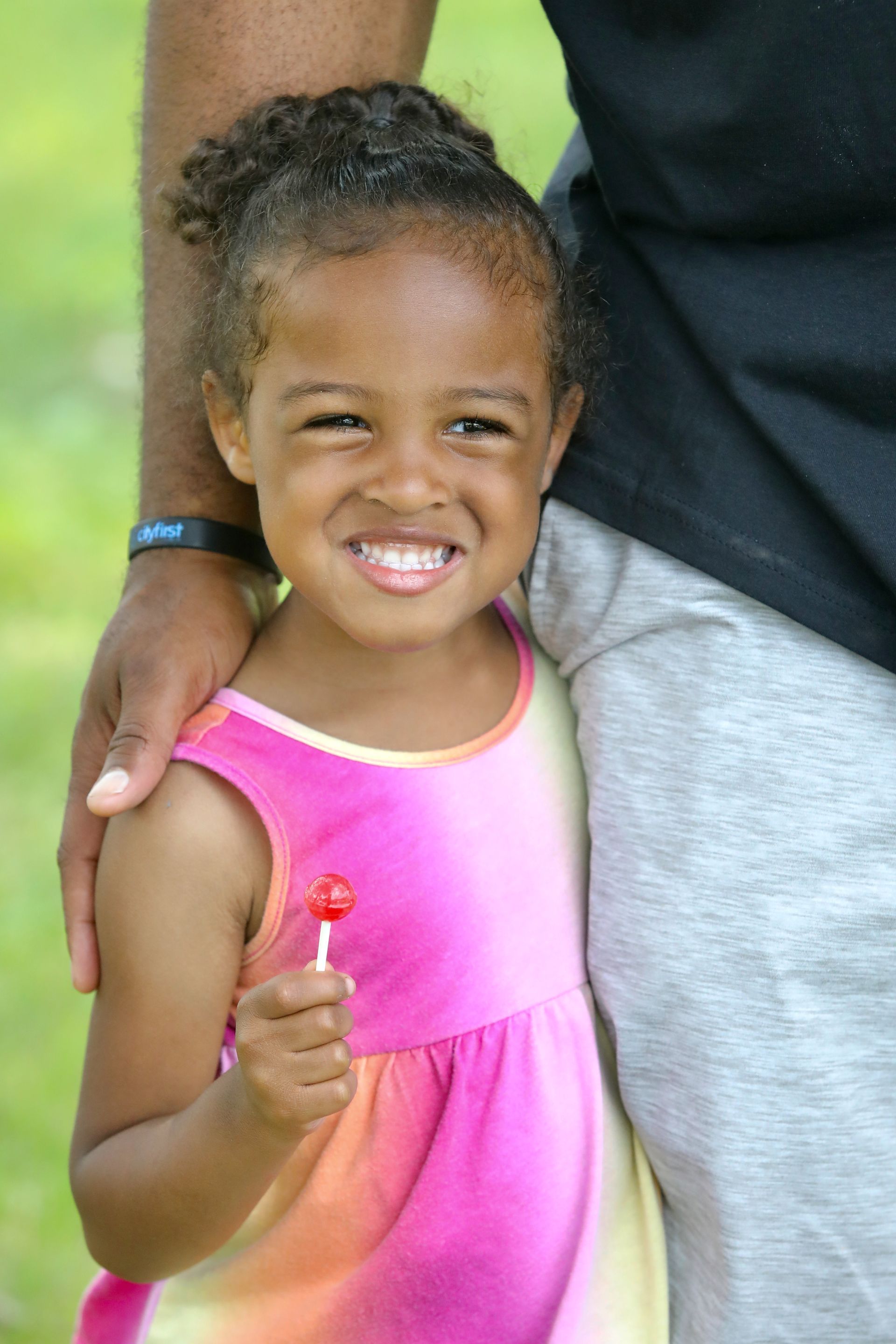 Girl holding lollipop, smiling next to an adult's arm in a park.