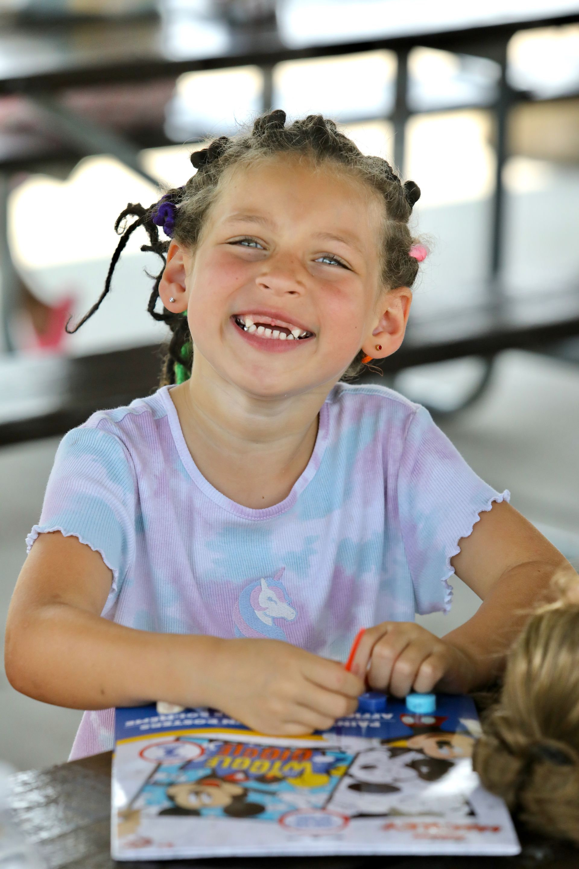 Girl with dreadlocks smiles, missing front teeth, at a table with a craft kit.