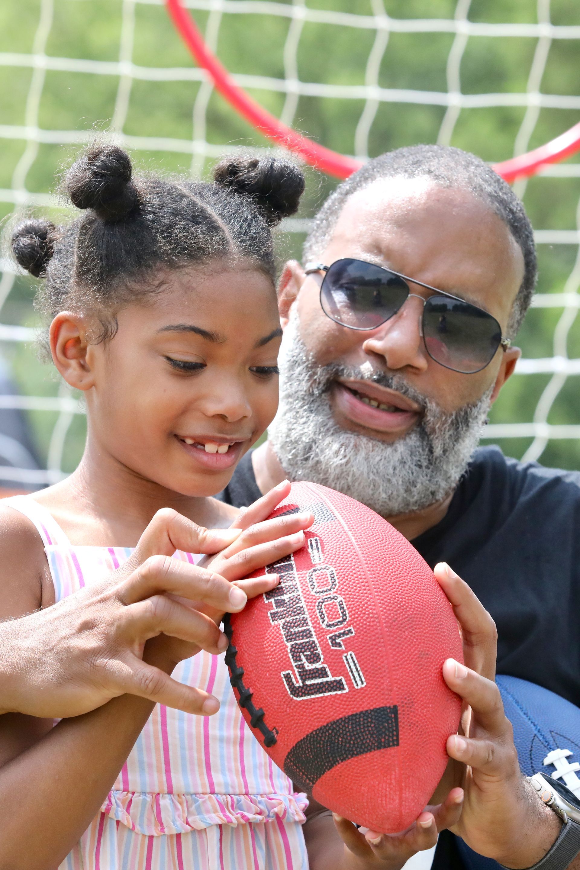 Man and young girl hold a red football, smiling. A green mesh fence is in the background.