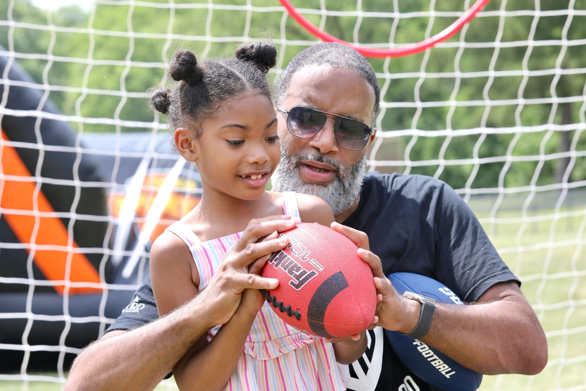 Man and girl holding a football in front of a sports net; sunny outdoor setting.