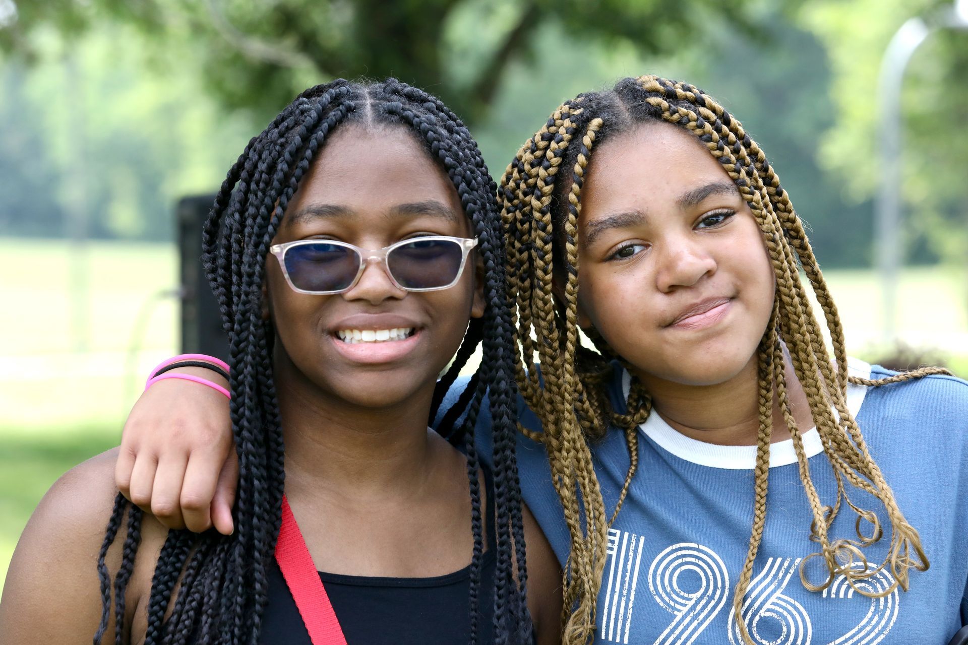 Two people with braided hair smiling, one wearing sunglasses, arms around each other, outdoors.