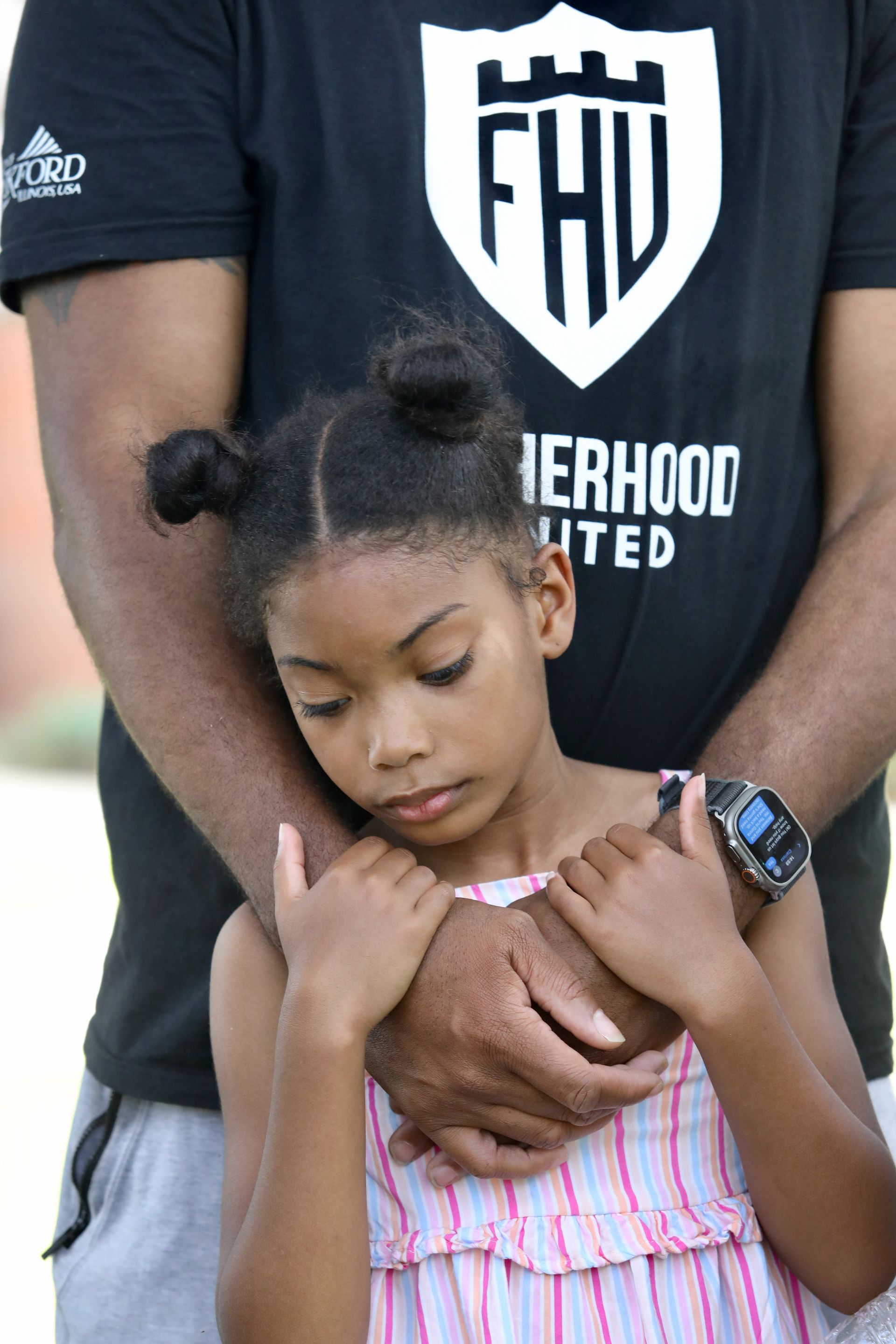 Man hugging a child, both looking down. Man wears a black shirt with a white logo.