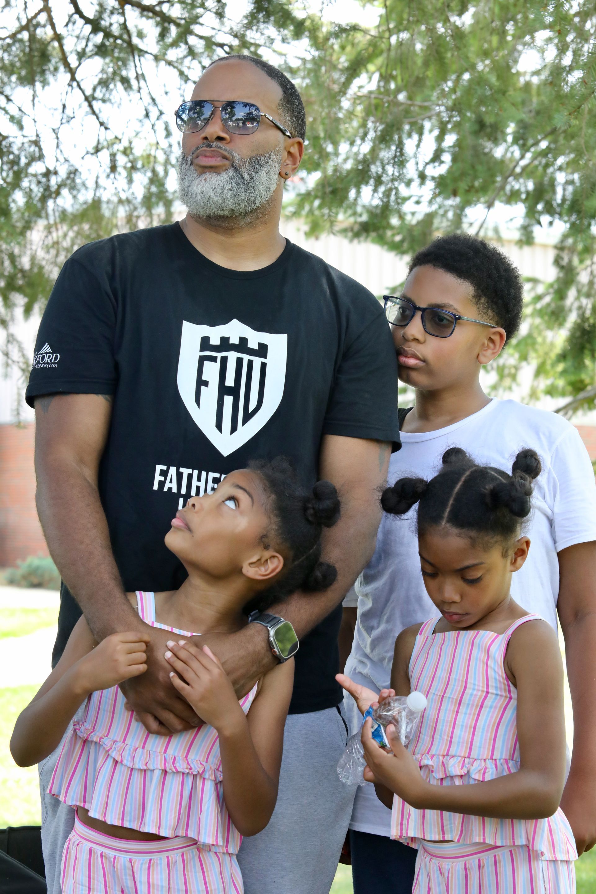 Man with three children outdoors, looking upwards. Children wear sunglasses; two girls have their hair up.