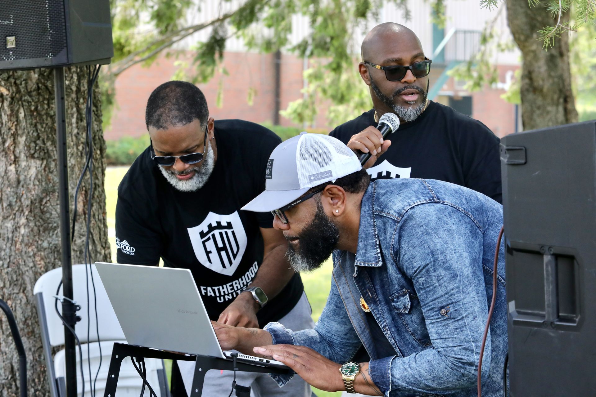 Three men with sunglasses at an outdoor event, using a laptop, microphone, and speakers.