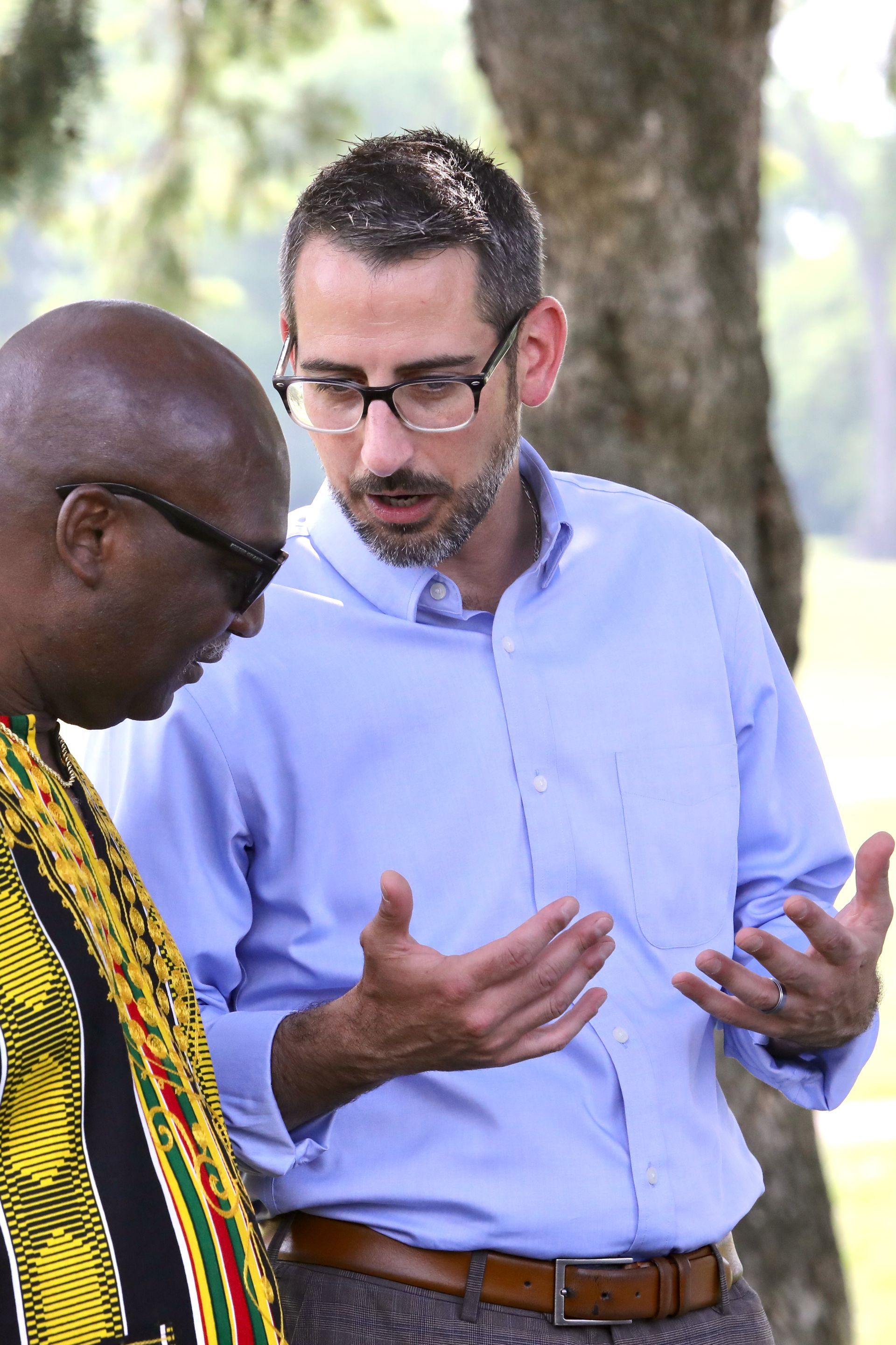 Two men outdoors, one in a patterned shirt, the other in a blue shirt, gesturing and talking, focused.