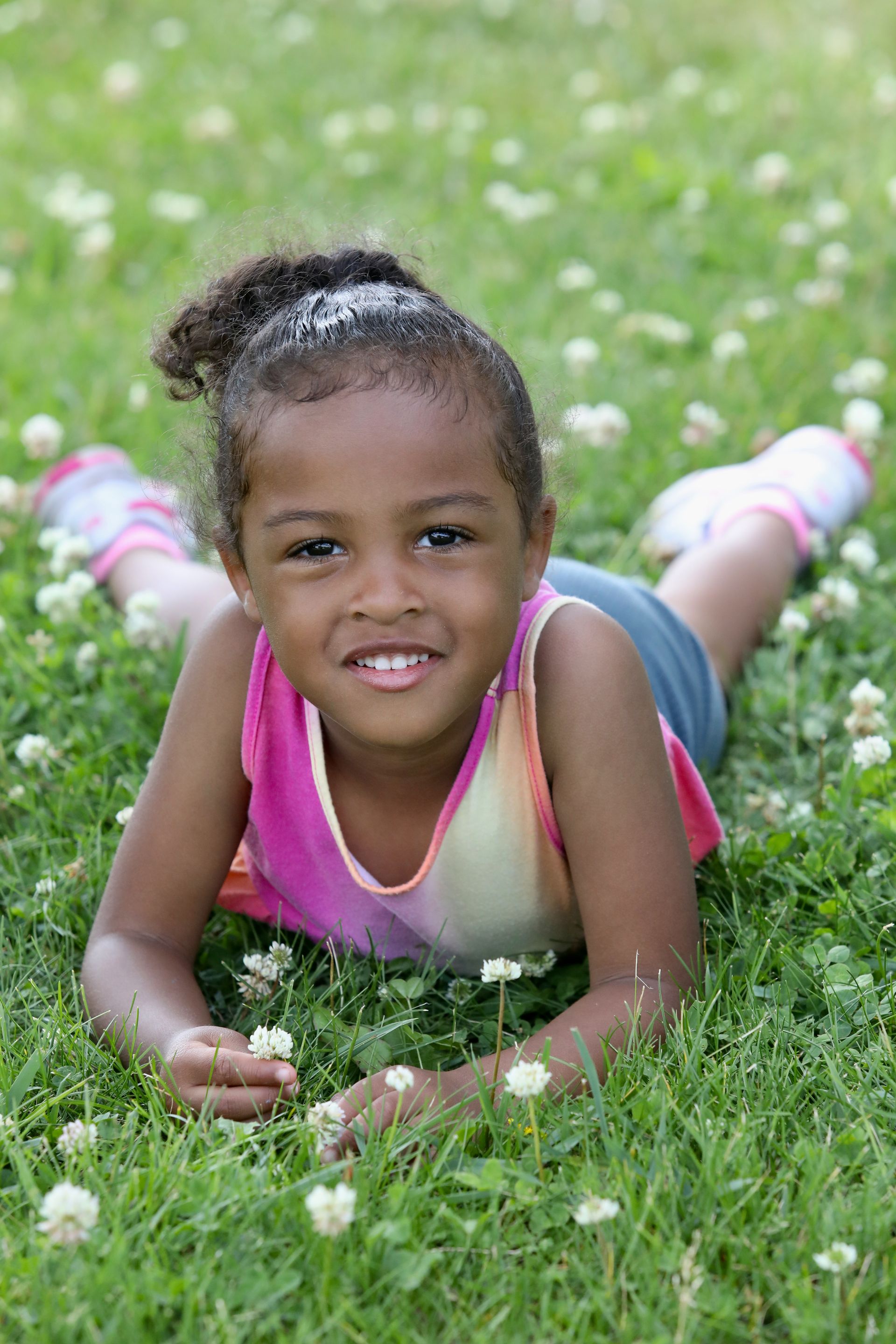 Girl with dark skin lying on grass, smiling. Wearing colorful top and shorts, in a field of small white flowers.