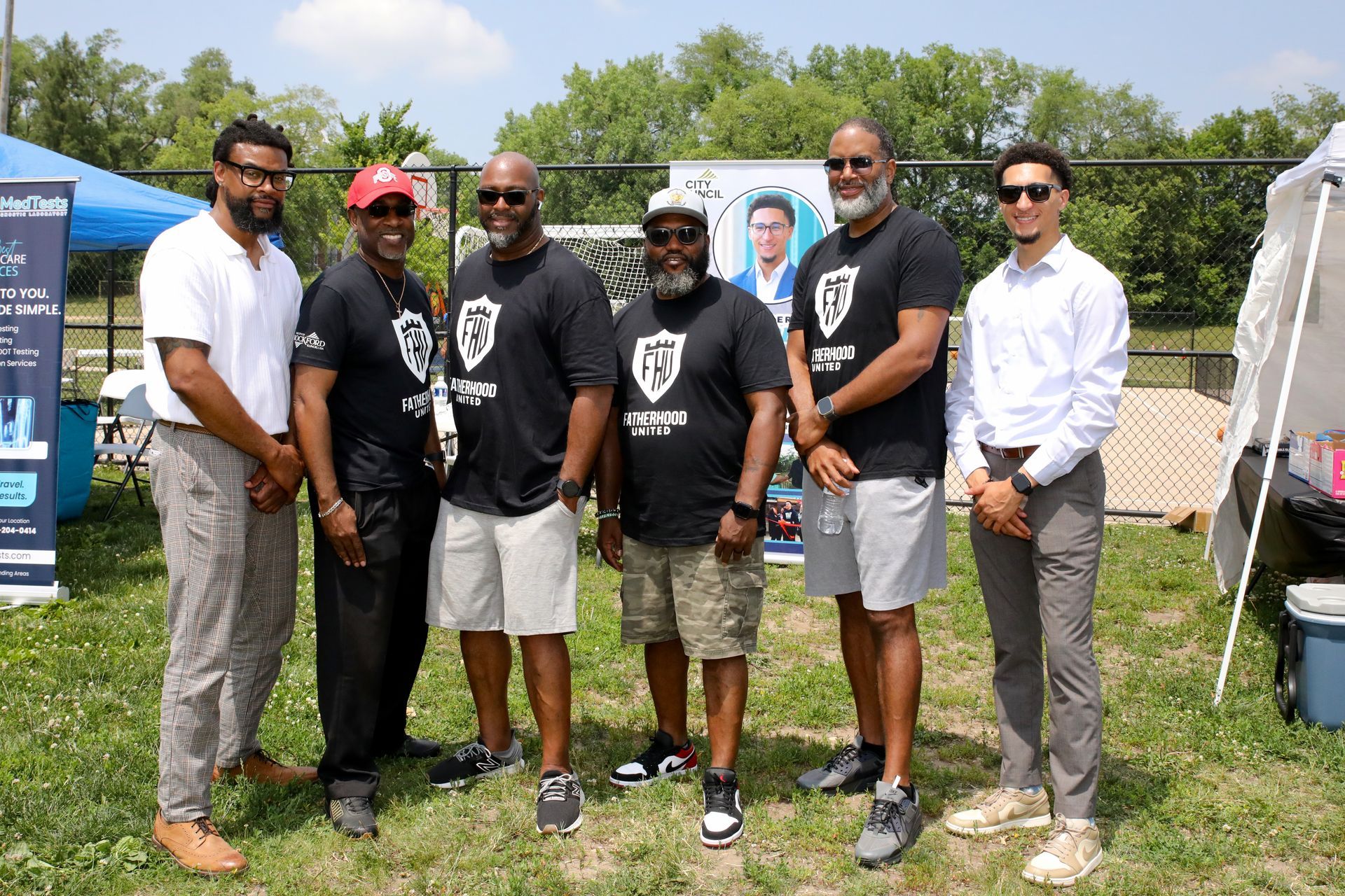 Six men standing in front of booths on a sunny day. They are wearing t-shirts and smiling.