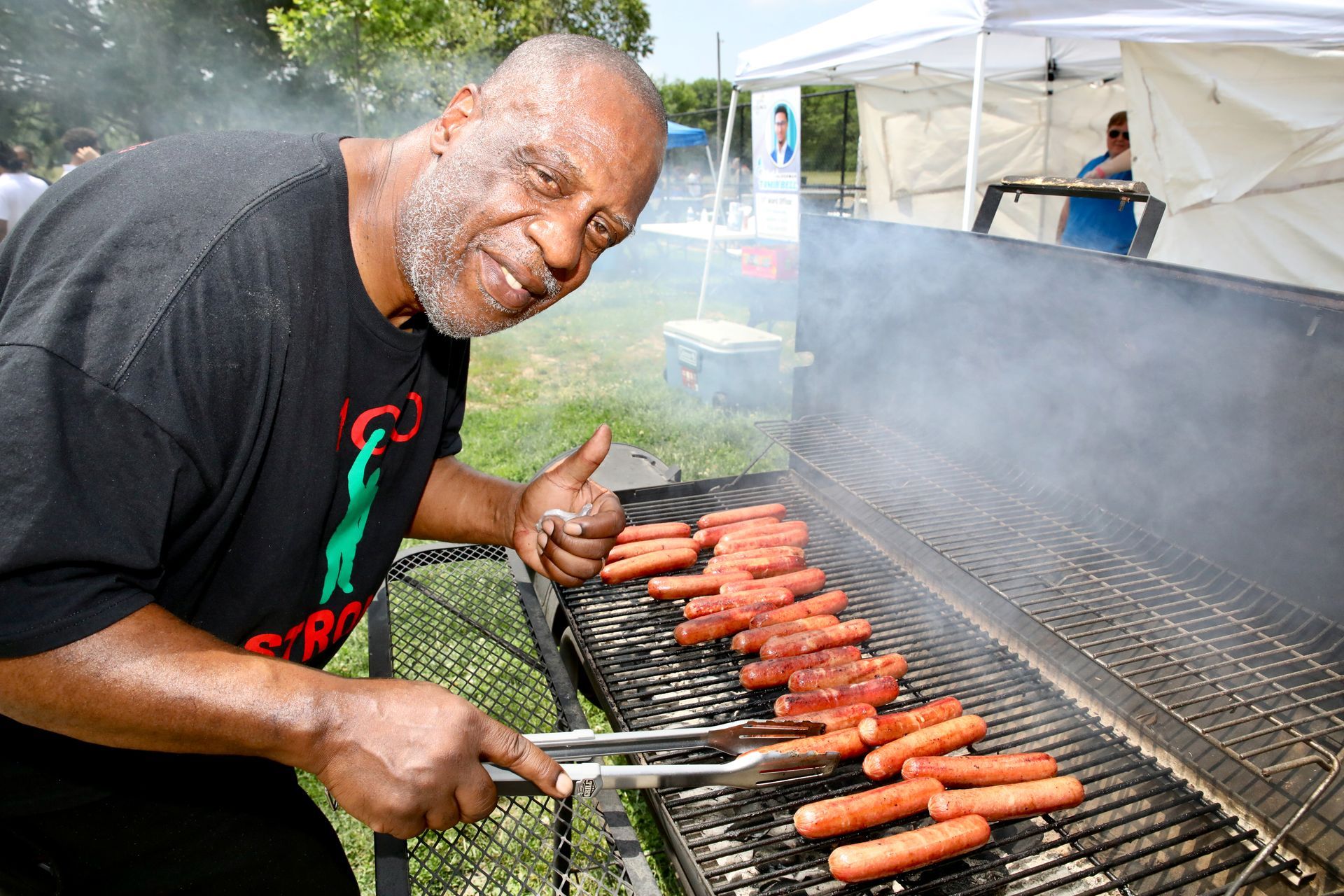 Man grilling hot dogs outdoors, giving a thumbs up.