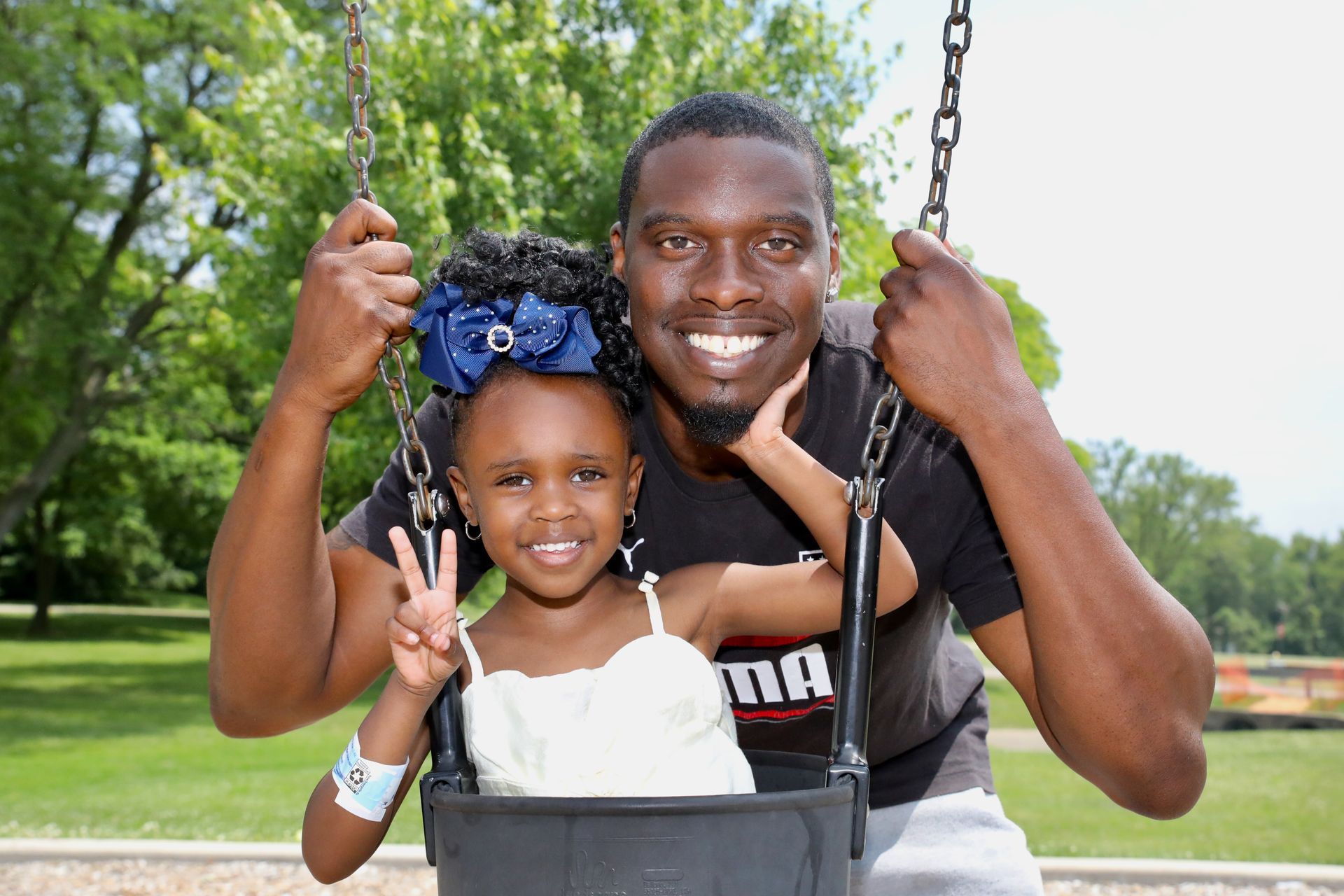 Father and child smiling on a swing set. The child is giving a peace sign. They are outdoors.