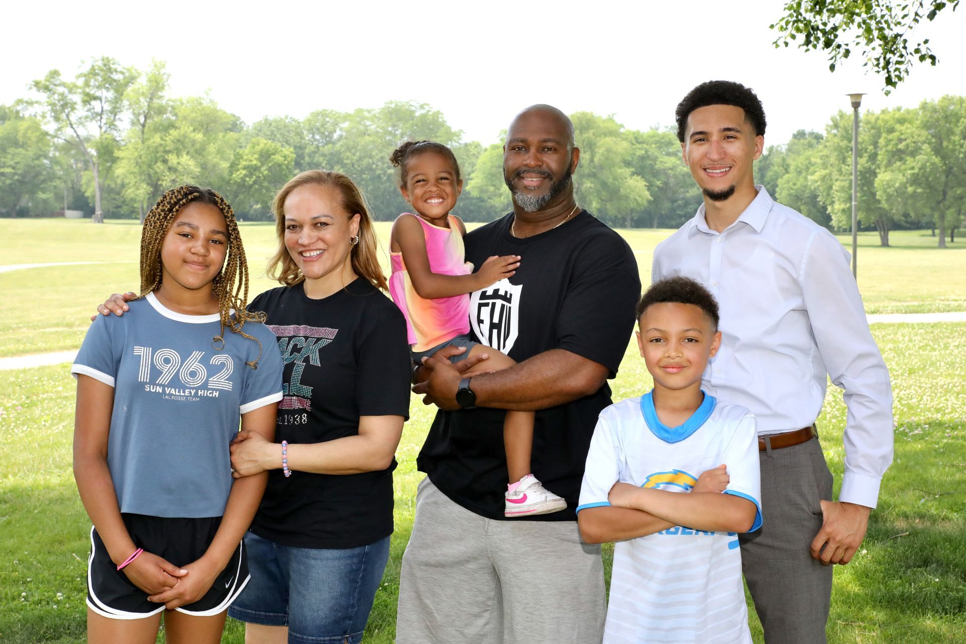 Family of six smiling in a park; woman holding a toddler, other children pose beside them.