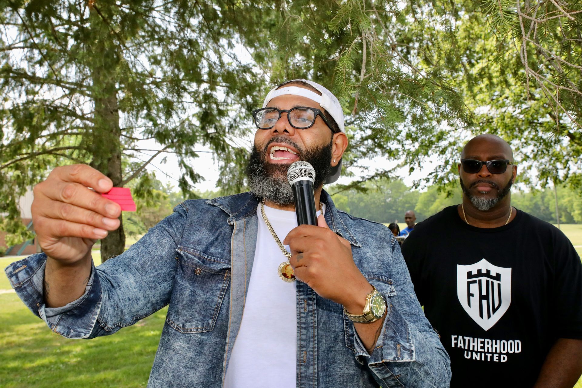 Man with beard and microphone speaking outdoors, another man in background.