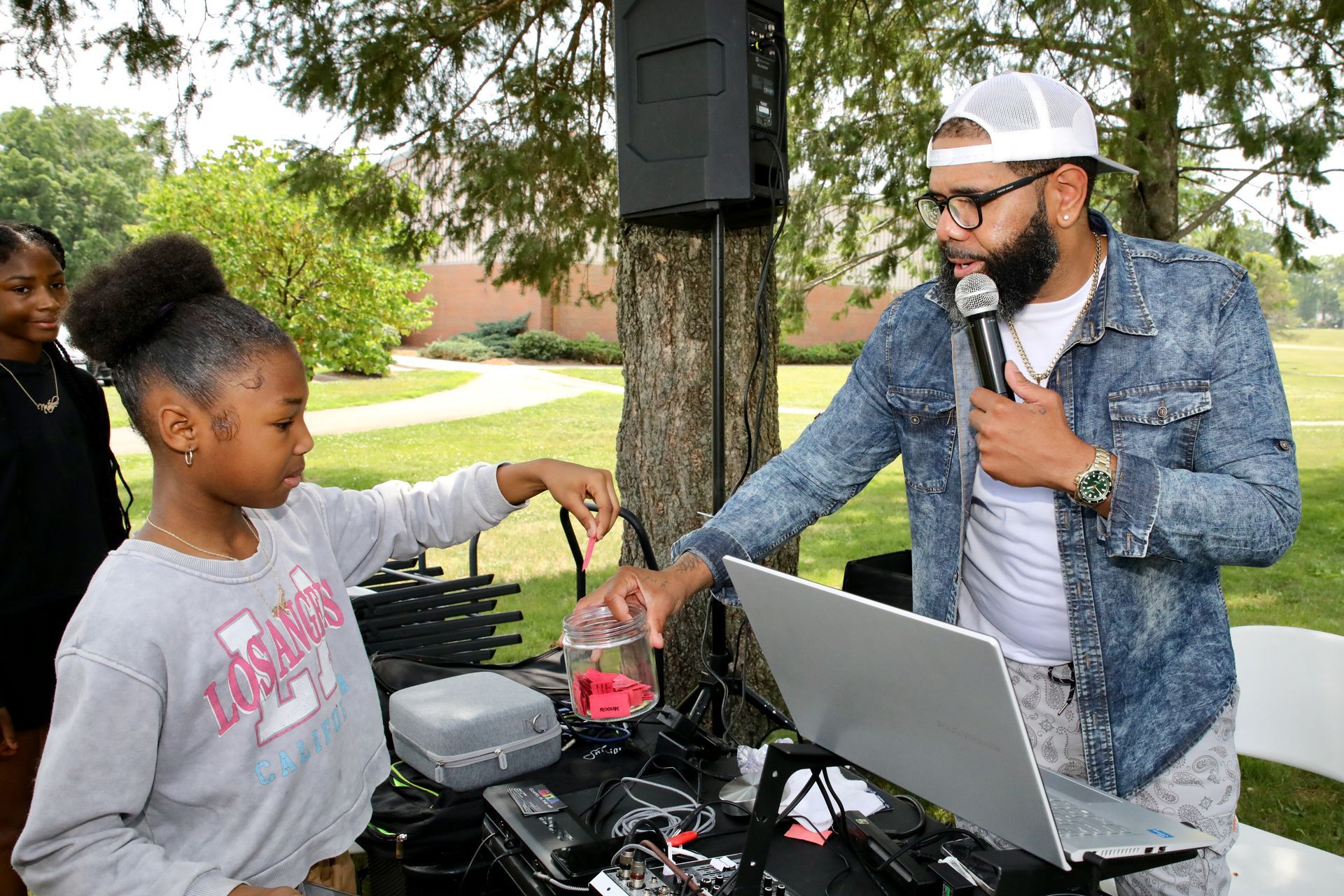 Man with microphone and young girl with a jar at an outdoor event. Laptop, speakers, trees visible.