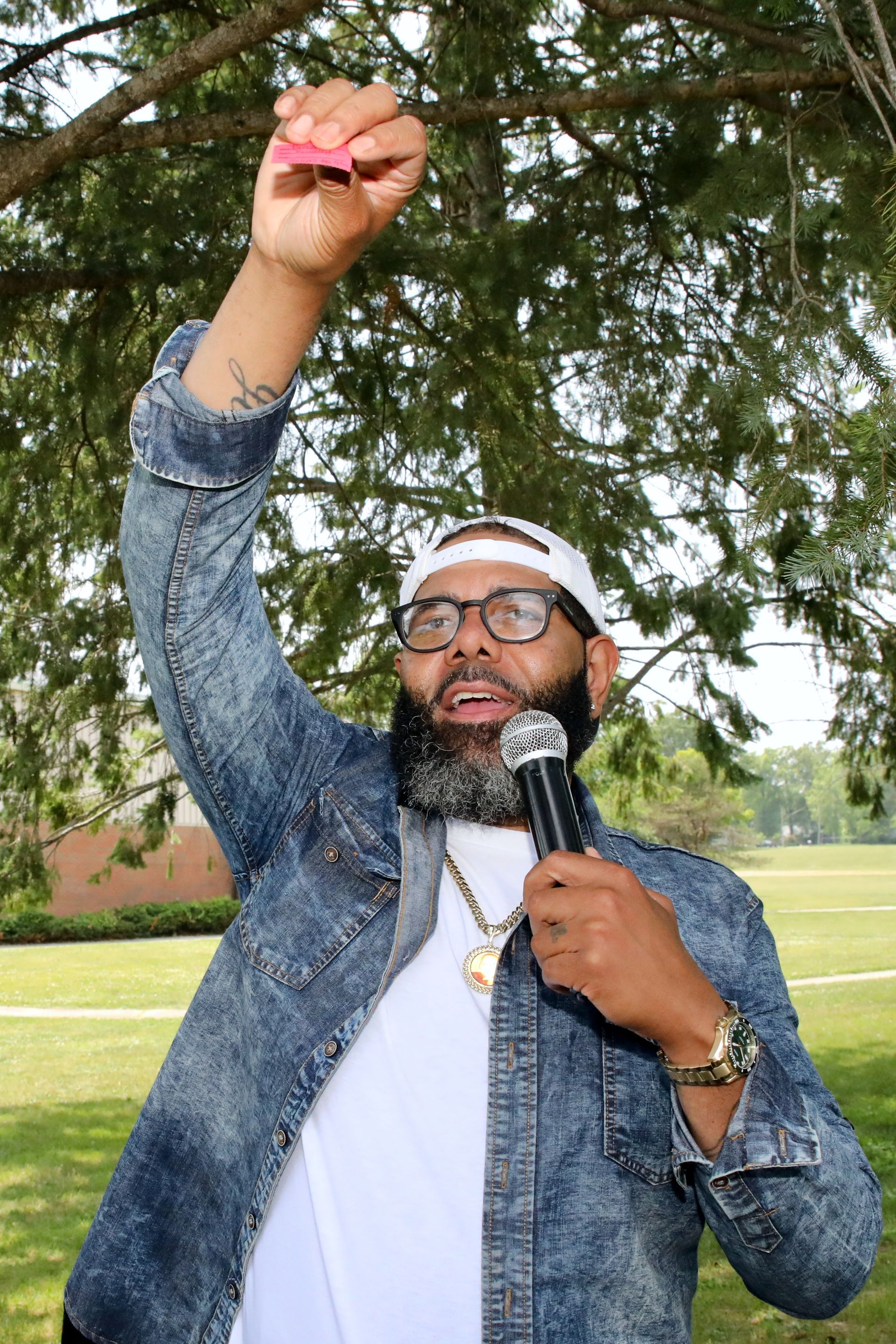 Man in denim jacket speaks into a microphone, holding up a pink item, outdoors.