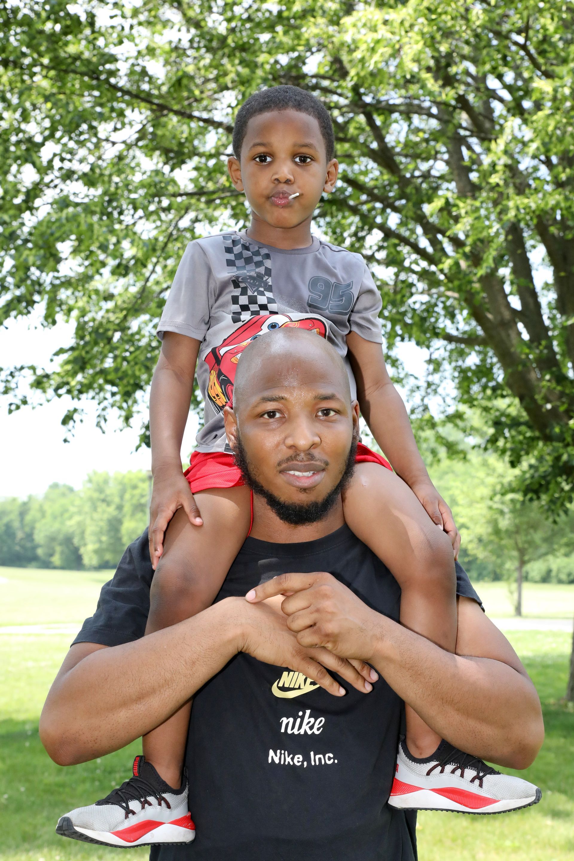 Man carries a child on his shoulders outdoors, both smiling. Green grass, trees in the background.