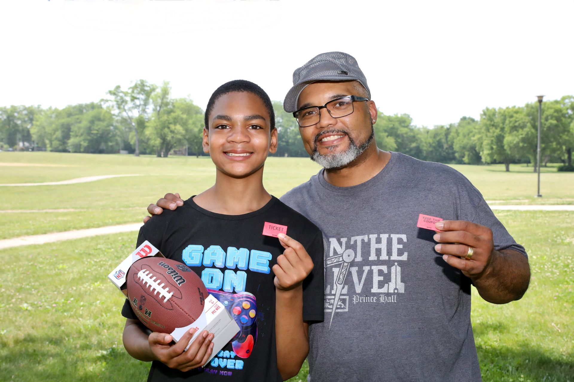 Man and boy pose outdoors holding football and game tickets, smiling.