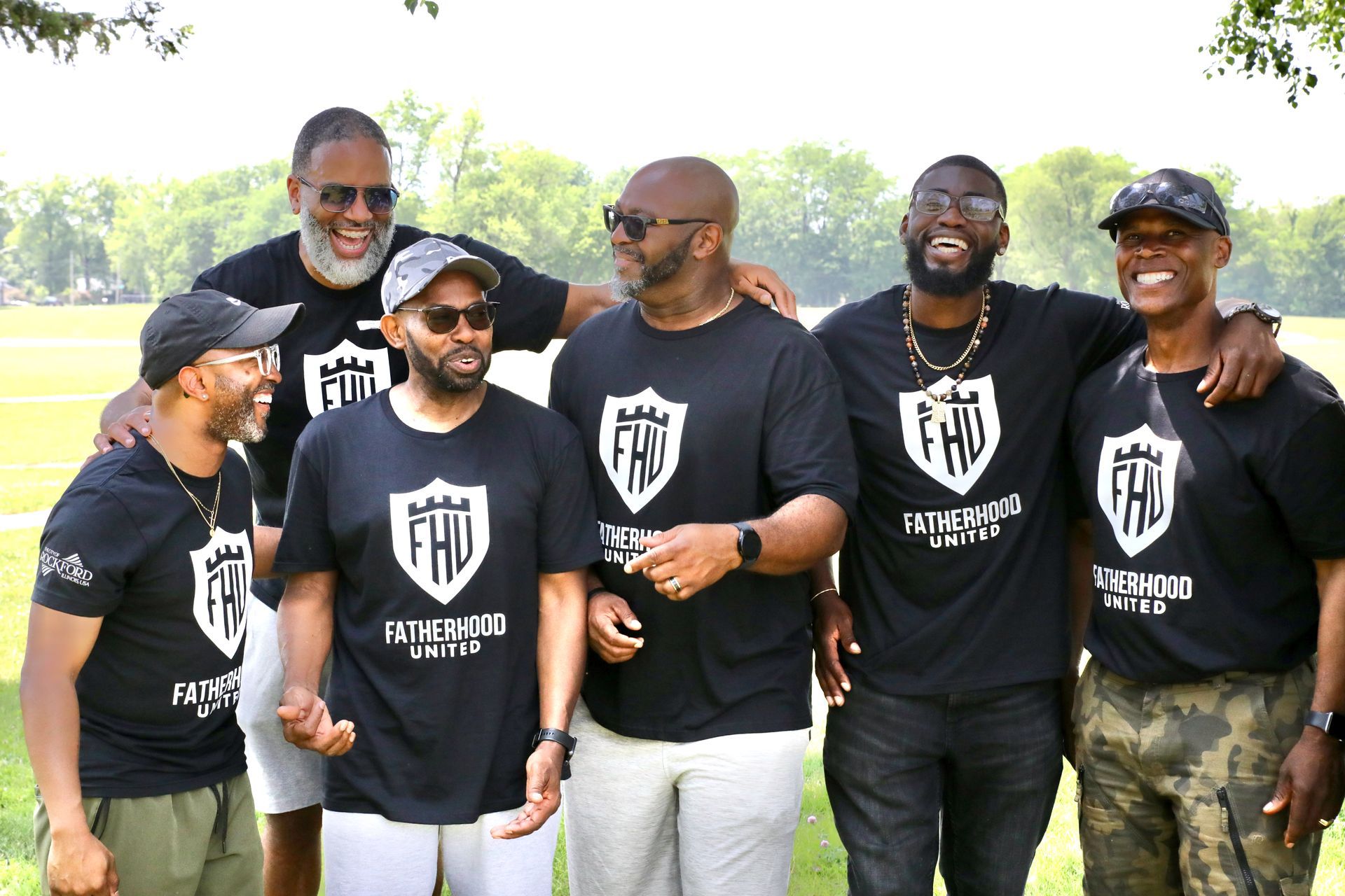 Six men in black shirts with logo, laughing and embracing outdoors.