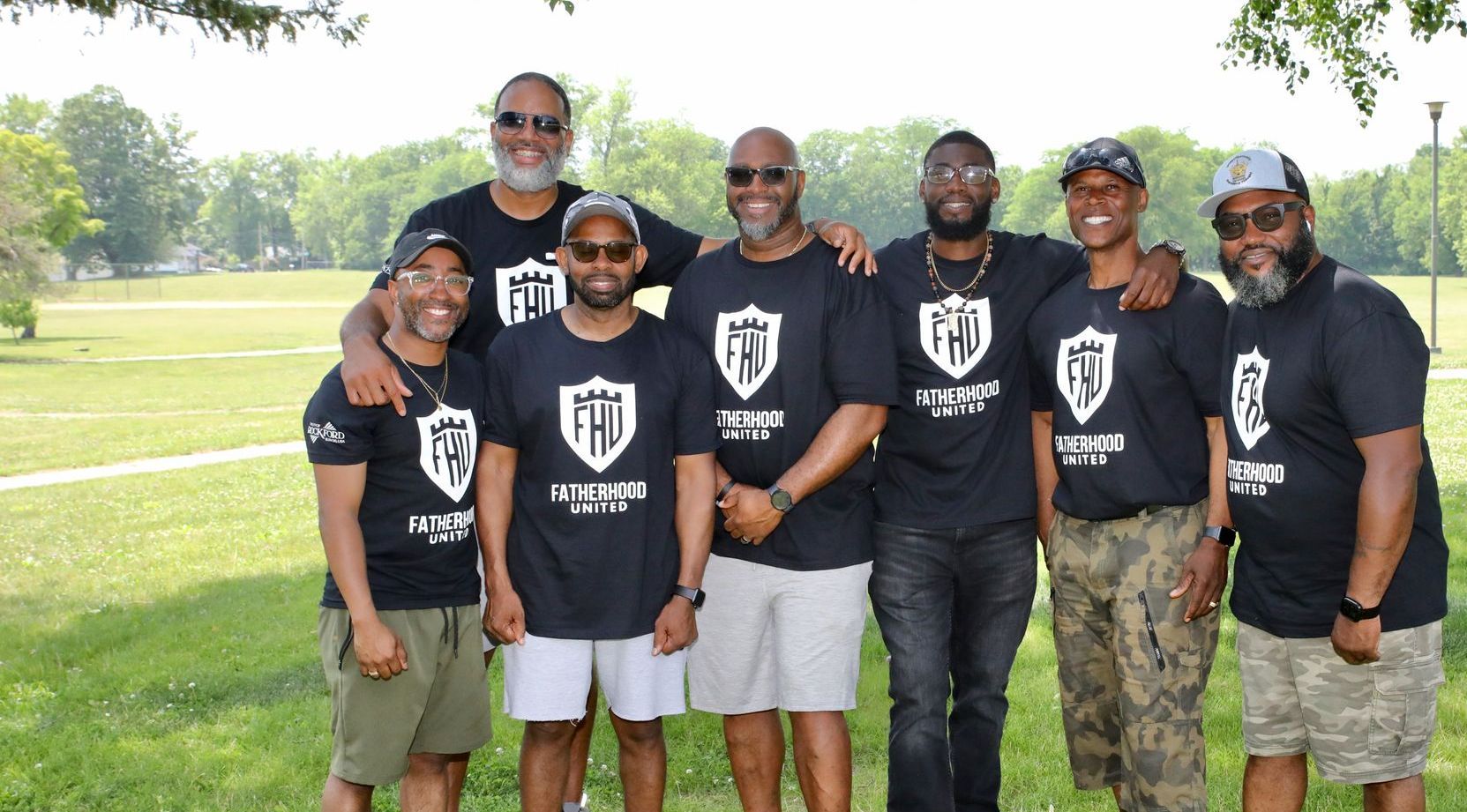 Seven men in matching black shirts pose together outdoors, arms around each other.