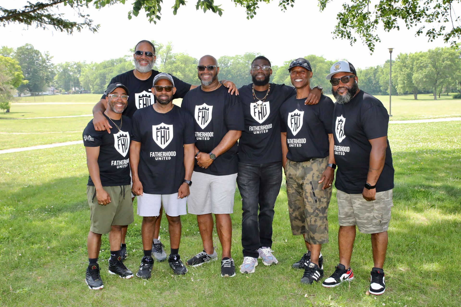 Group of men in black shirts with a logo, standing together outdoors on a sunny day.